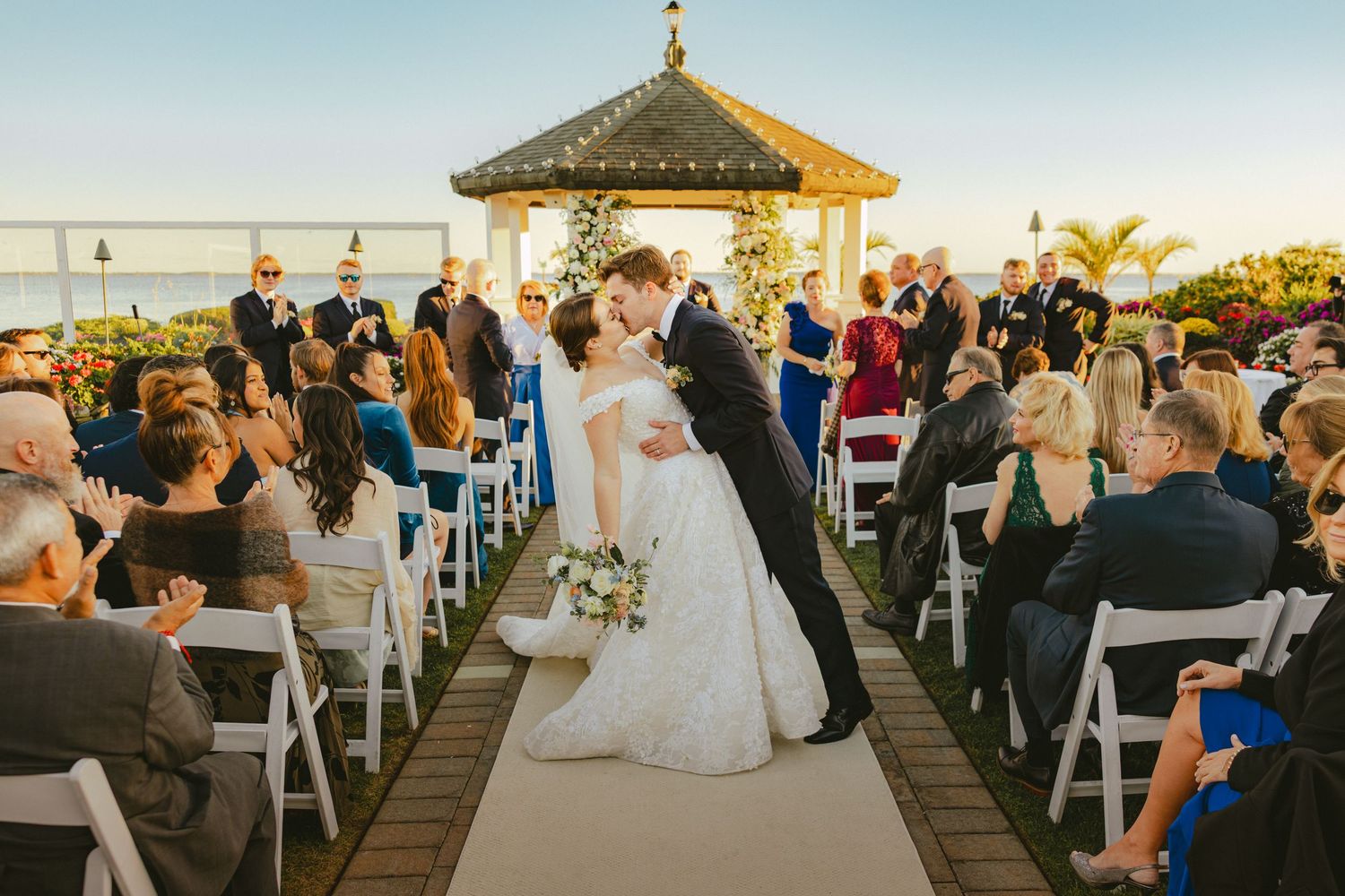 Bride and groom share first kiss at outdoor rooftop gazebo ceremony with seated guests watching.