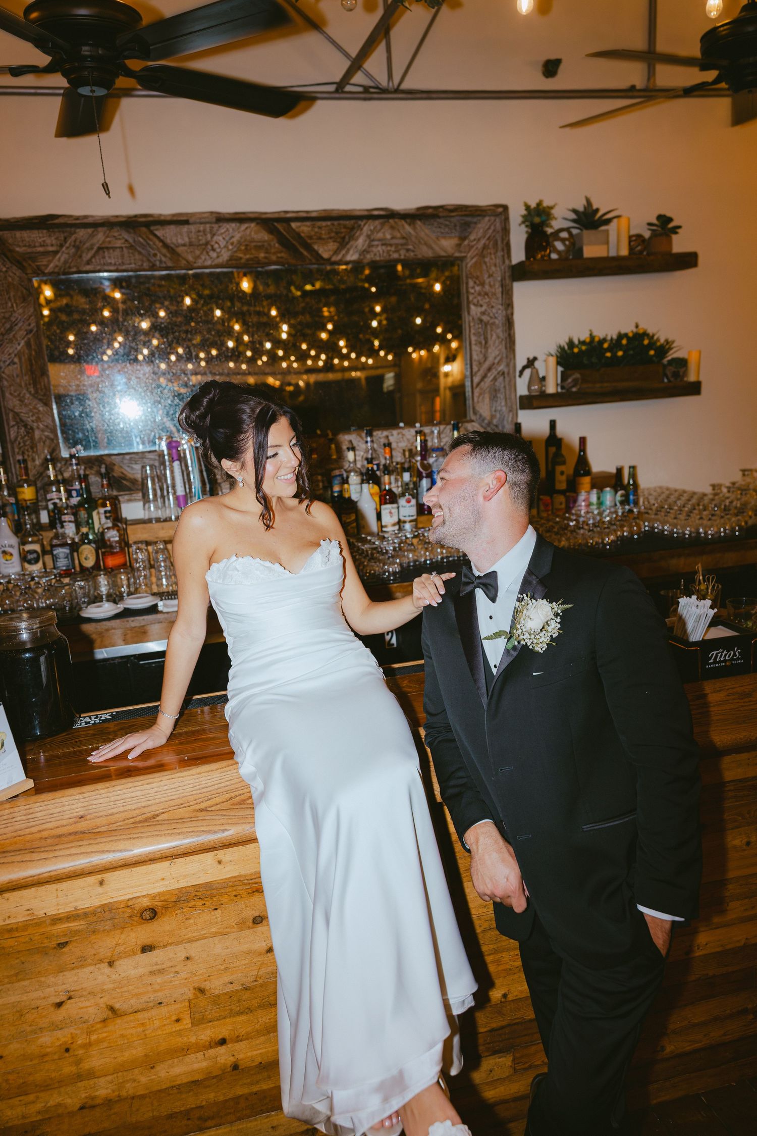 Bride and groom share a playful moment together at a rustic bar with string lights.