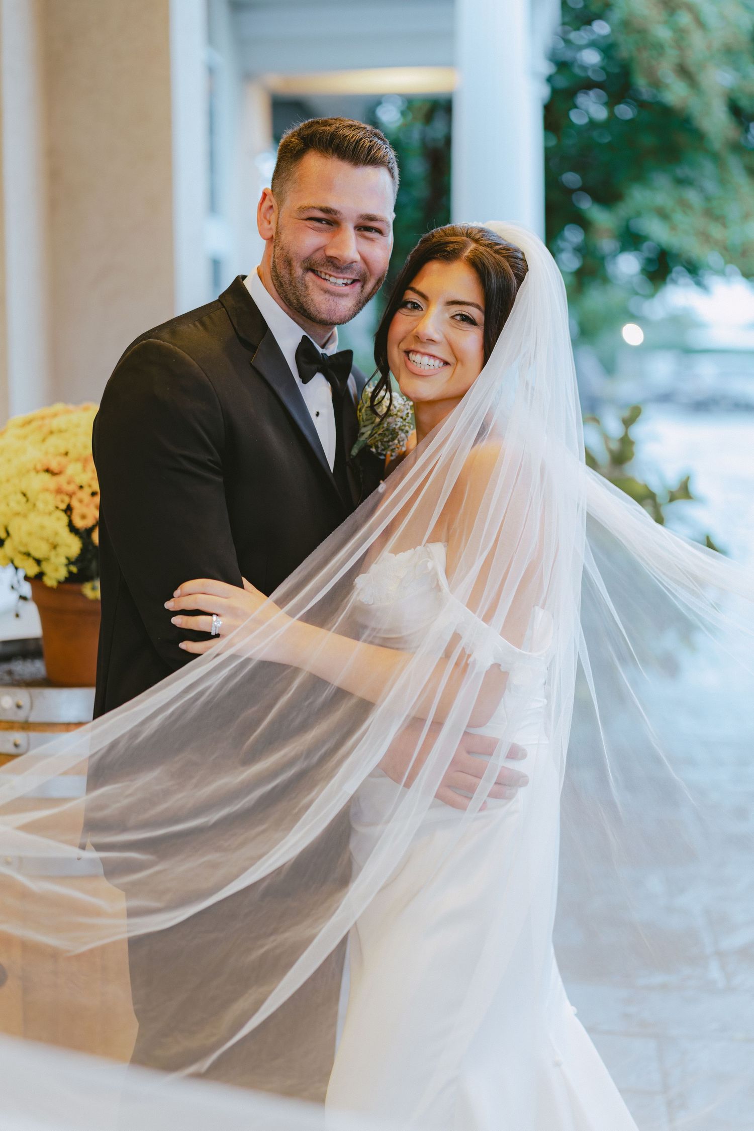 Smiling bride and groom pose outdoors with flowing veil billowing around them after wedding.