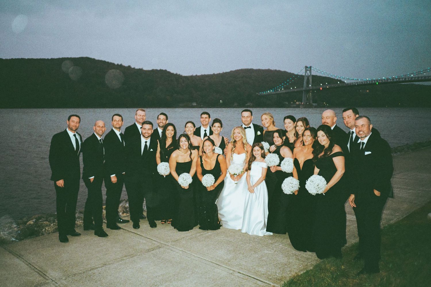 Large wedding party in black formal attire posing by a river with a bridge at dusk.