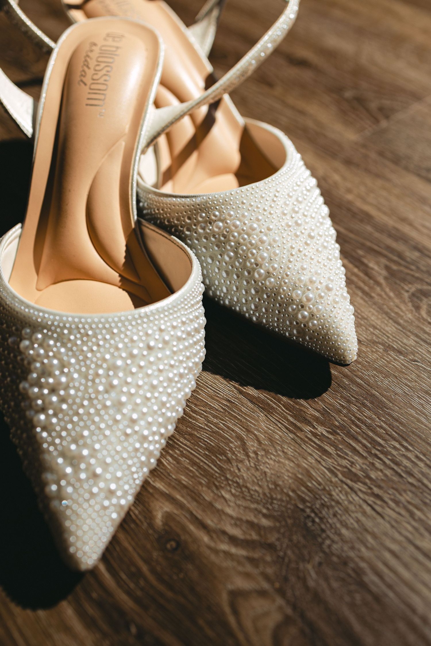 Pearl-embellished pointed toe bridal heels with ankle straps displayed on wooden floor in sunlight.