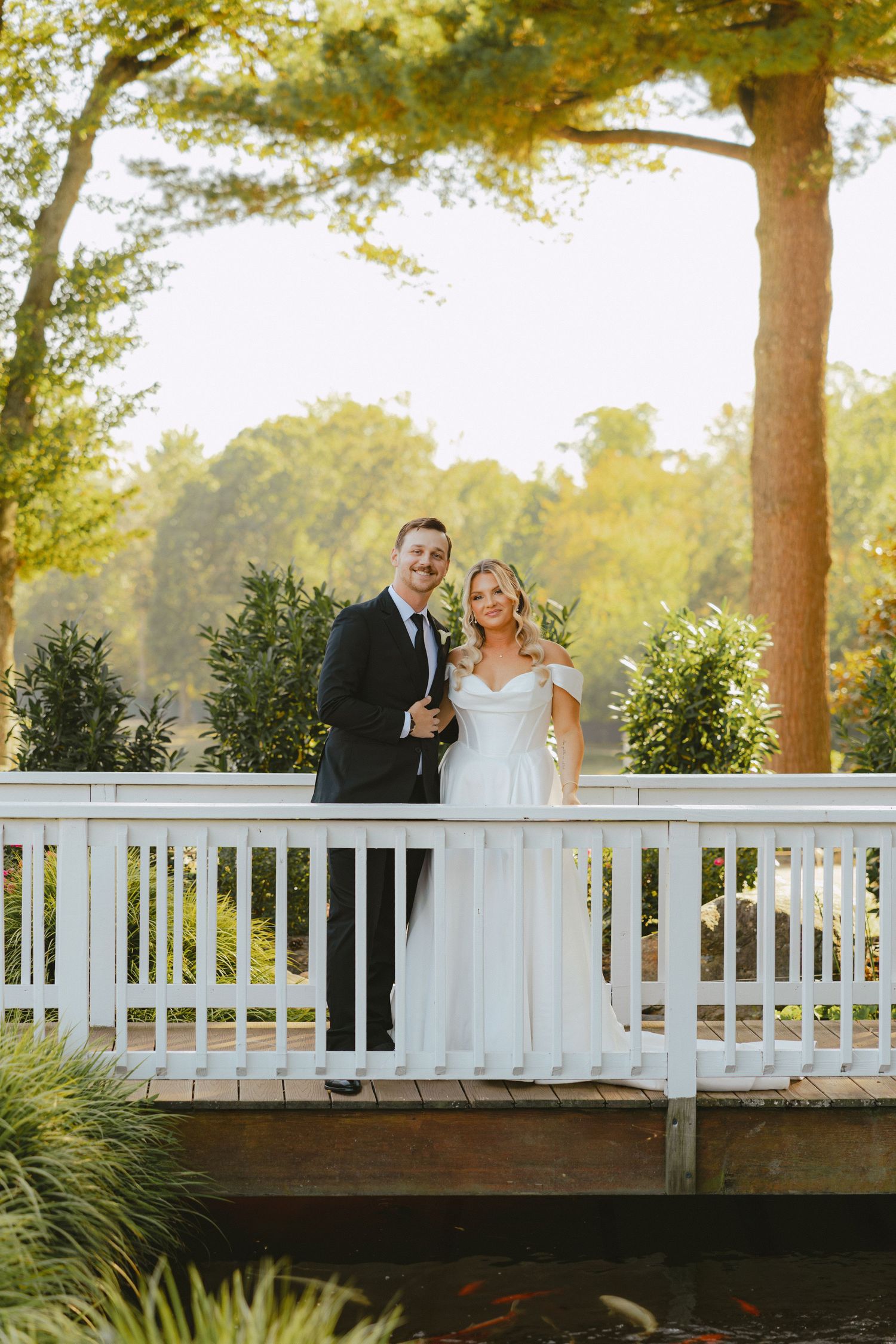 Smiling bride and groom pose together on a white bridge overlooking a pond in a sunlit garden setting.