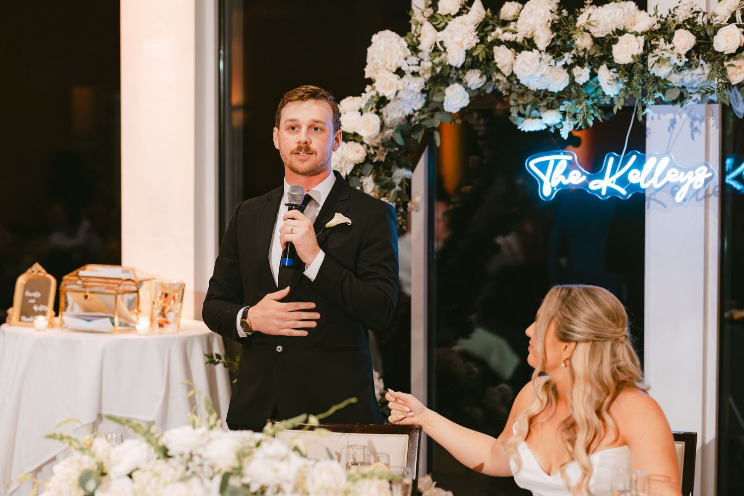 Groom speaks at wedding reception with microphone while bride listens near white floral decorations and neon sign.