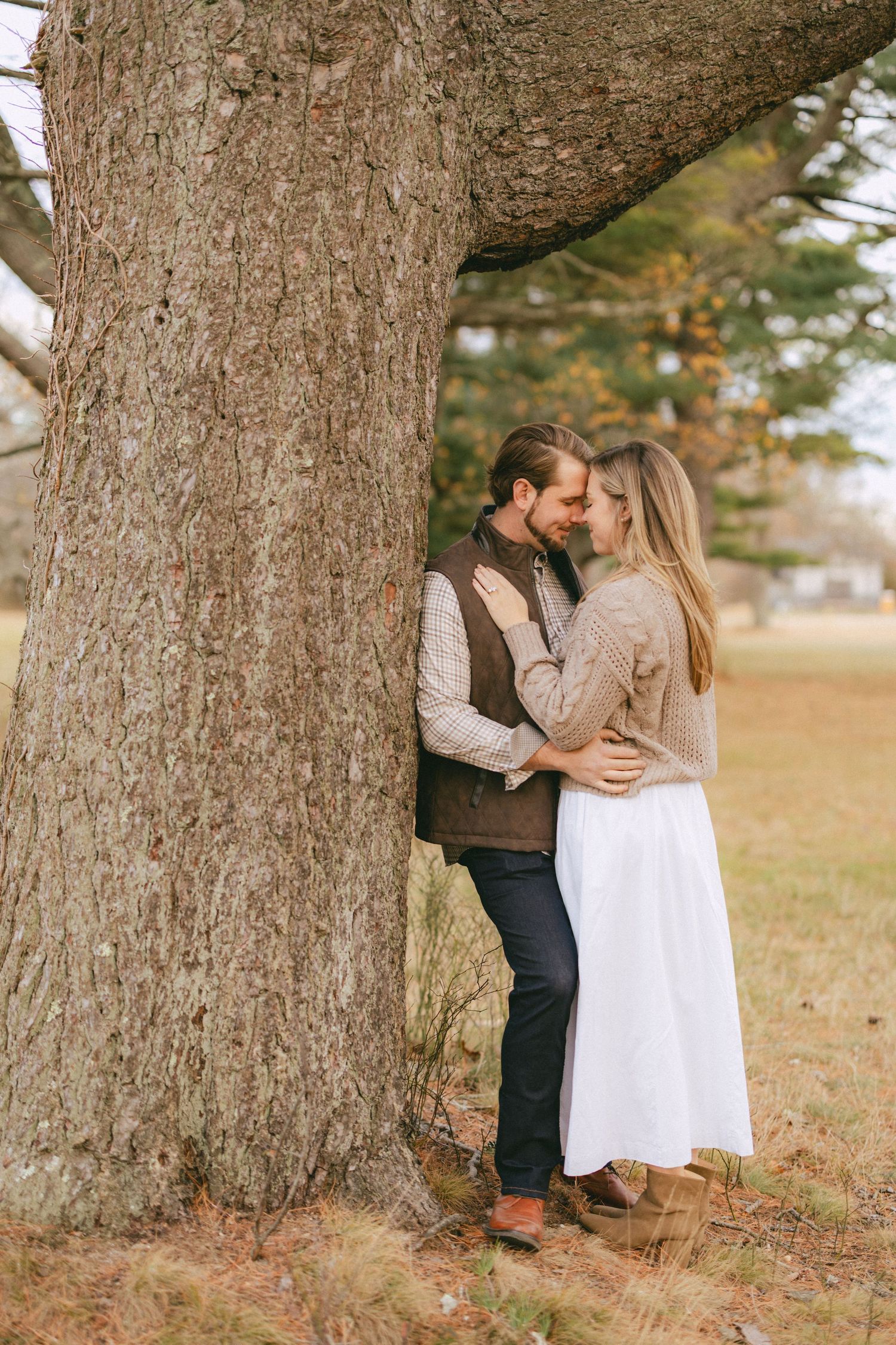 Couple embraces closely beside a large tree trunk in a peaceful autumn landscape.