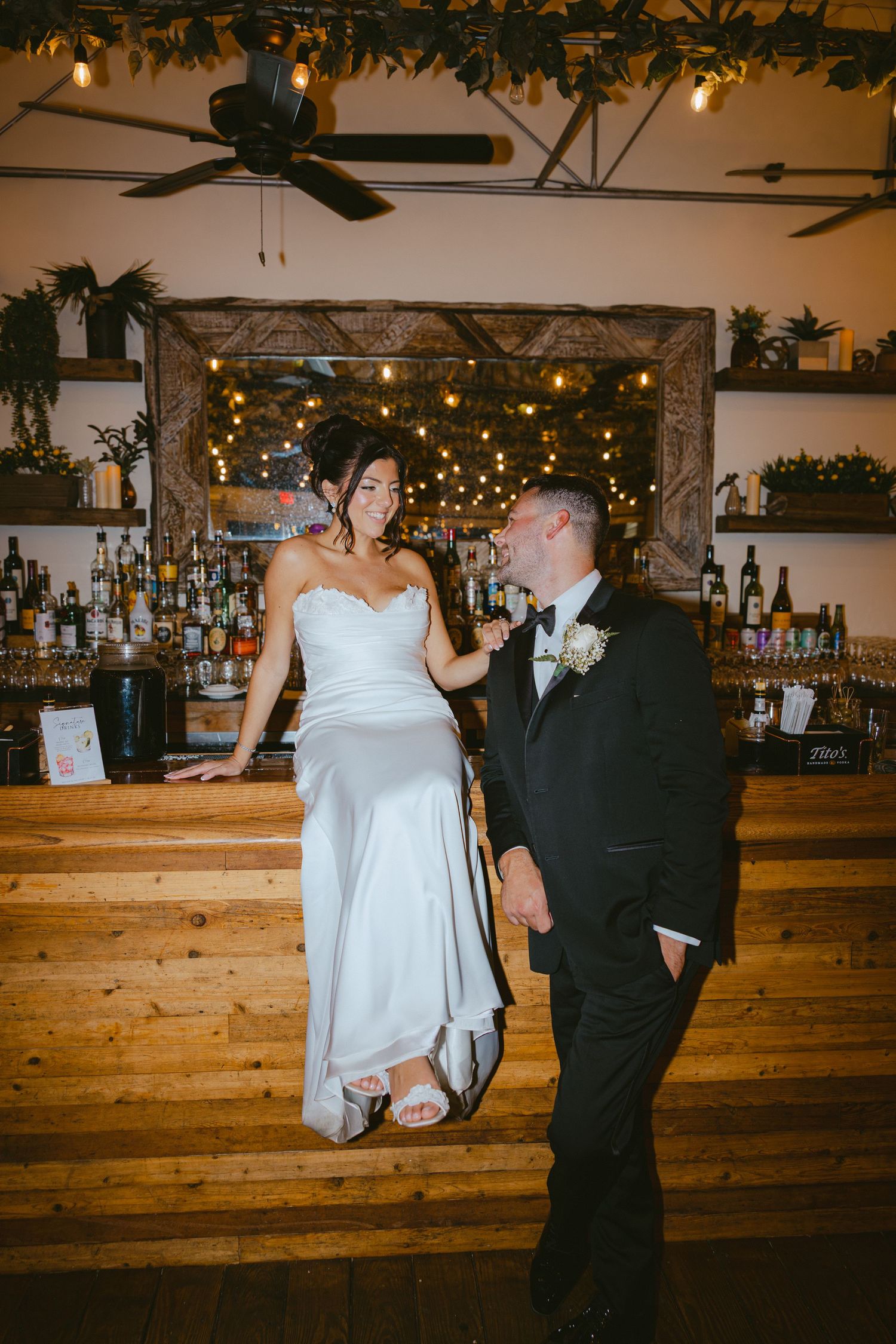 Bride and groom pose together at a rustic wooden bar with warm string lights.