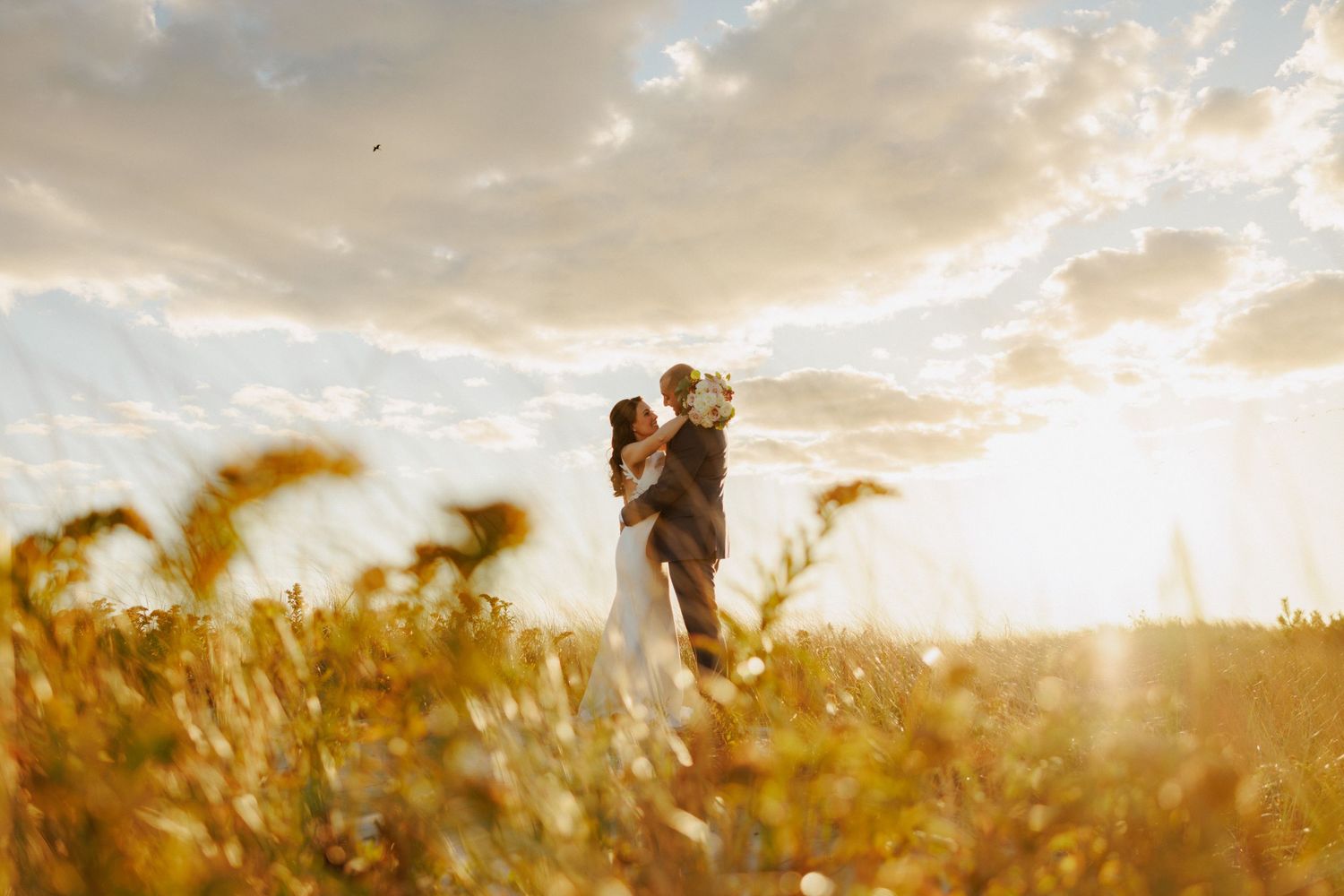 Bride and groom embrace in golden wheat field beneath dramatic cloudy sky at sunset.