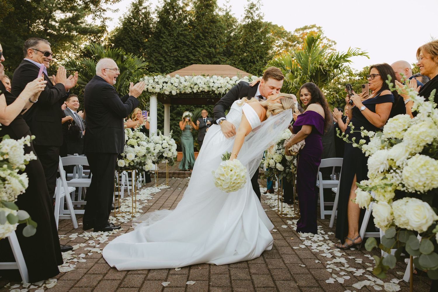 Bride and groom share romantic dip kiss in flower-lined aisle after outdoor wedding ceremony.