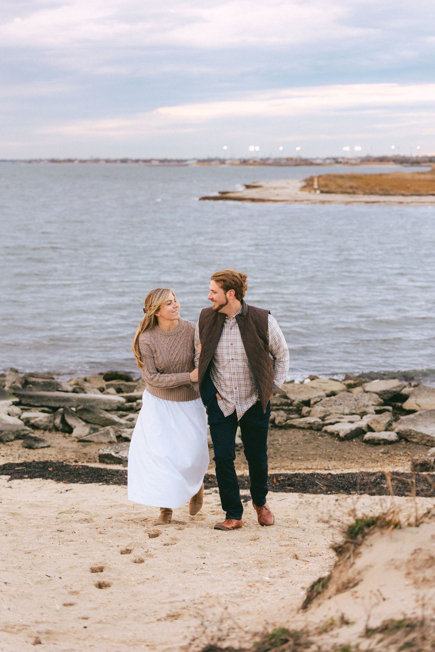 Happy couple strolls arm in arm along rocky sandy shoreline at sunset, smiling at each other lovingly.