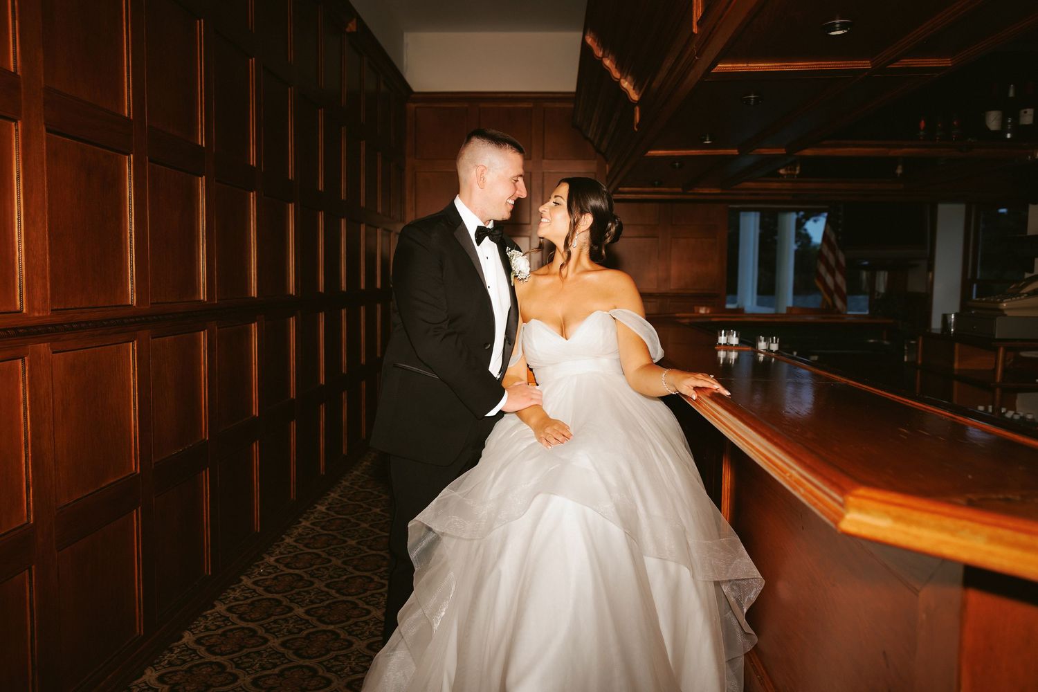 Bride in ball gown and groom in tuxedo sharing intimate moment at wooden bar.