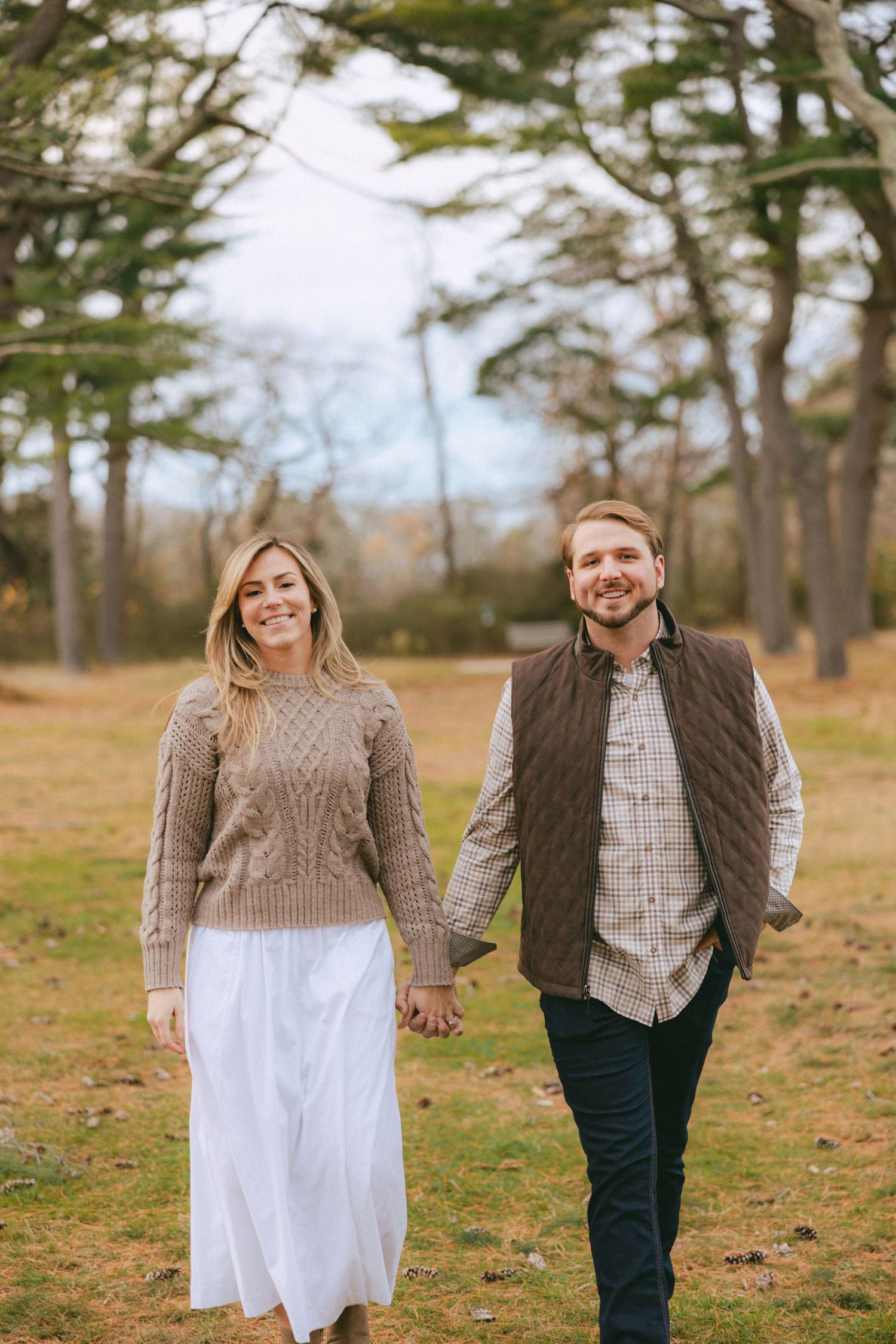 Smiling couple walks hand in hand through a scenic park with tall autumn trees.