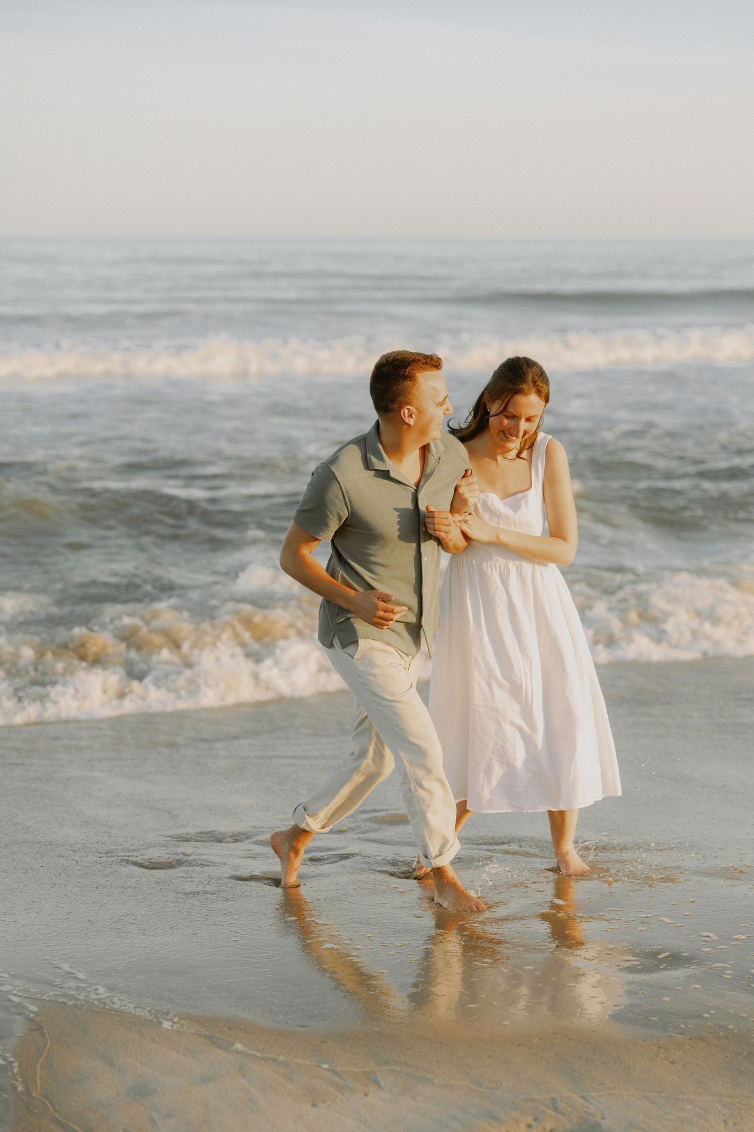 Laughing couple walking arm in arm along wet sandy beach shoreline with ocean waves behind.