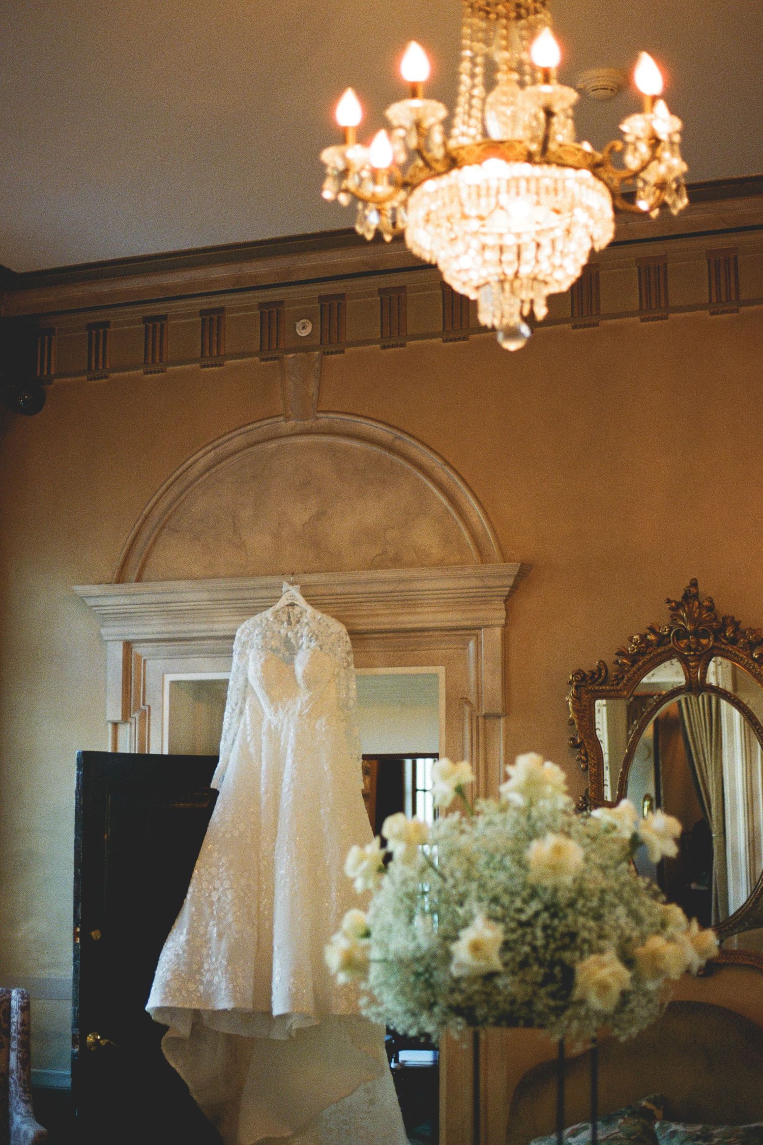 Lace wedding dress hanging under crystal chandelier in ornate room with white floral arrangement.