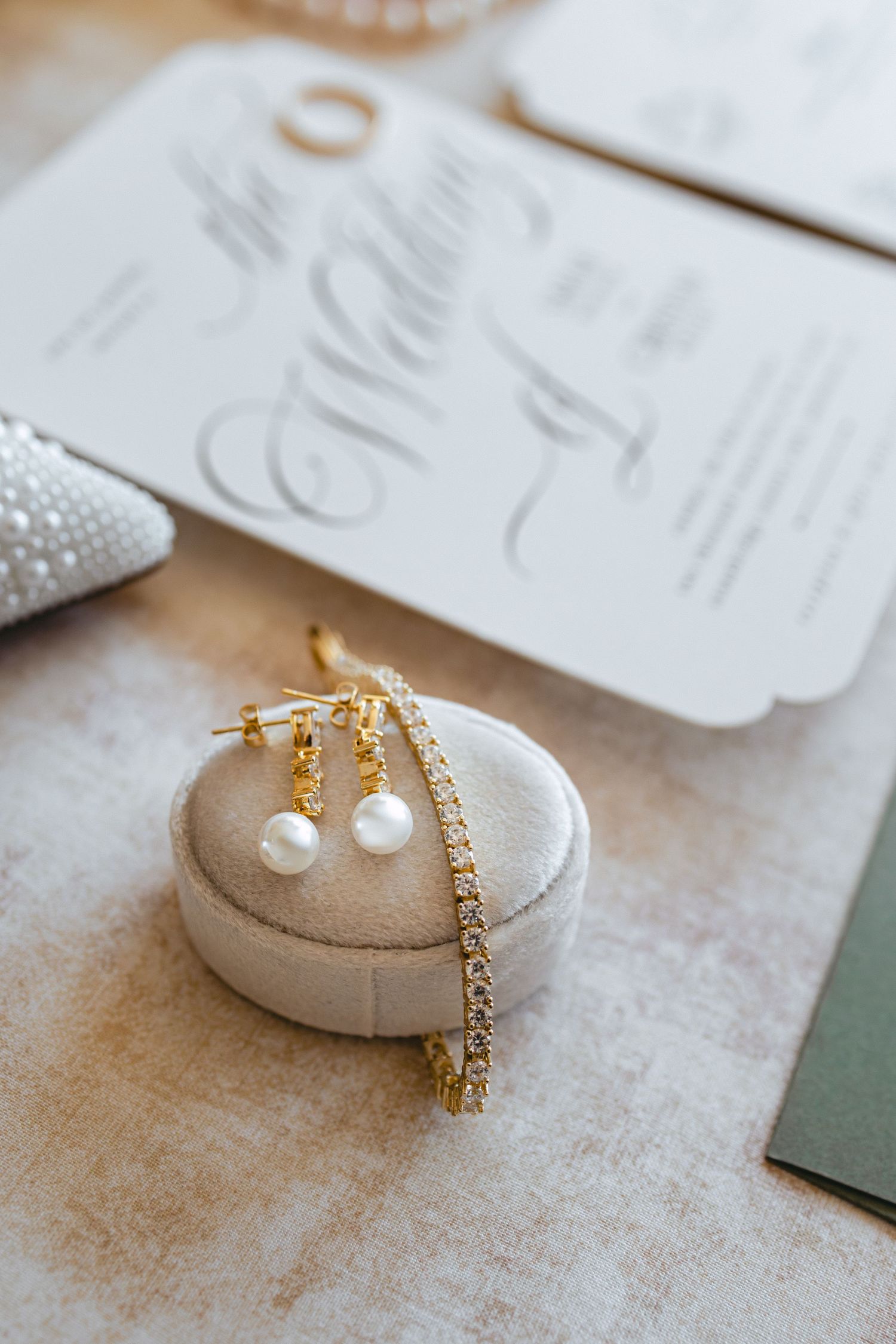 Gold pearl drop earrings and diamond tennis bracelet on velvet ring box with wedding invitation.