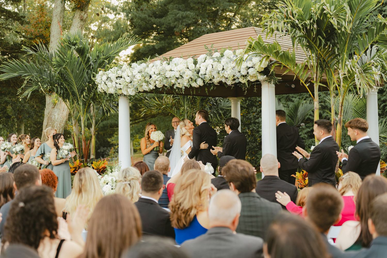 Guests watch bride and groom exchange vows under a floral gazebo at an outdoor ceremony.