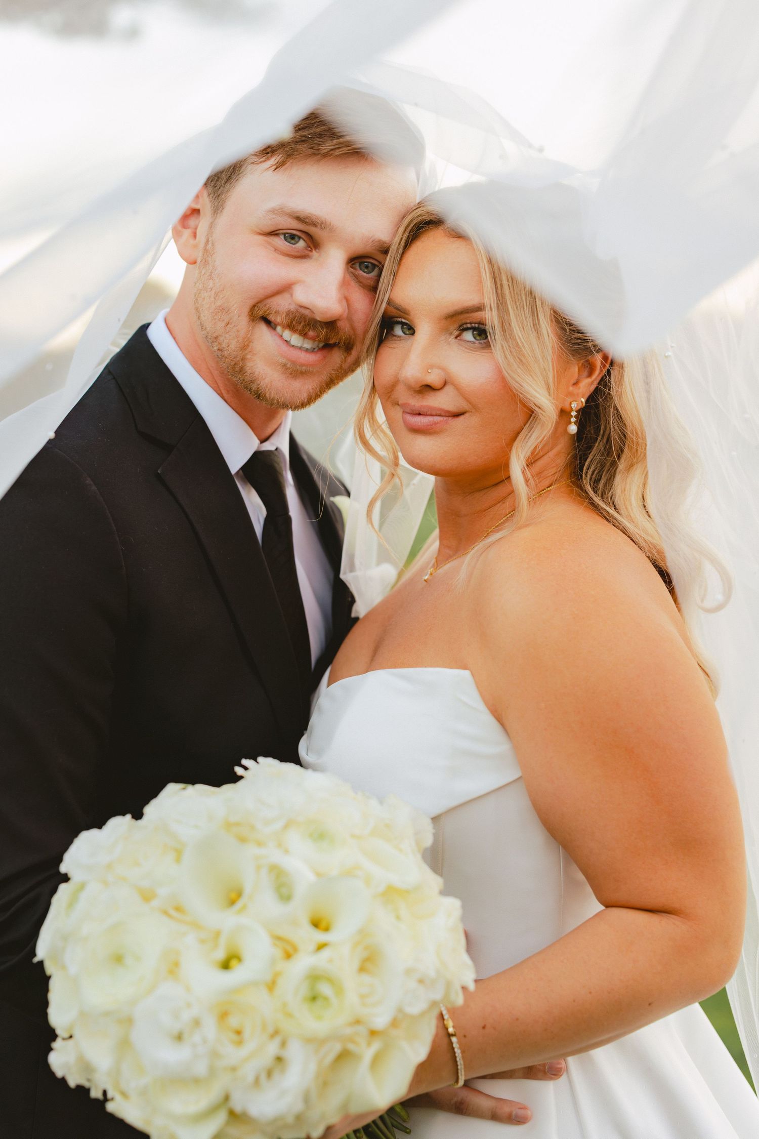 Smiling bride and groom pose together under a veil with a white rose bouquet.