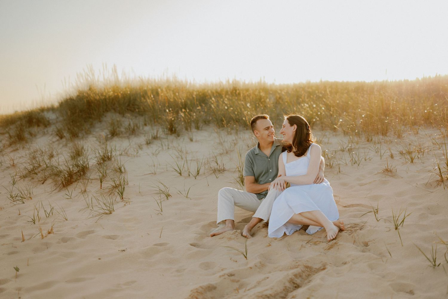 Engaged couple sits together kissing on sand dunes surrounded by beach grass at sunset.