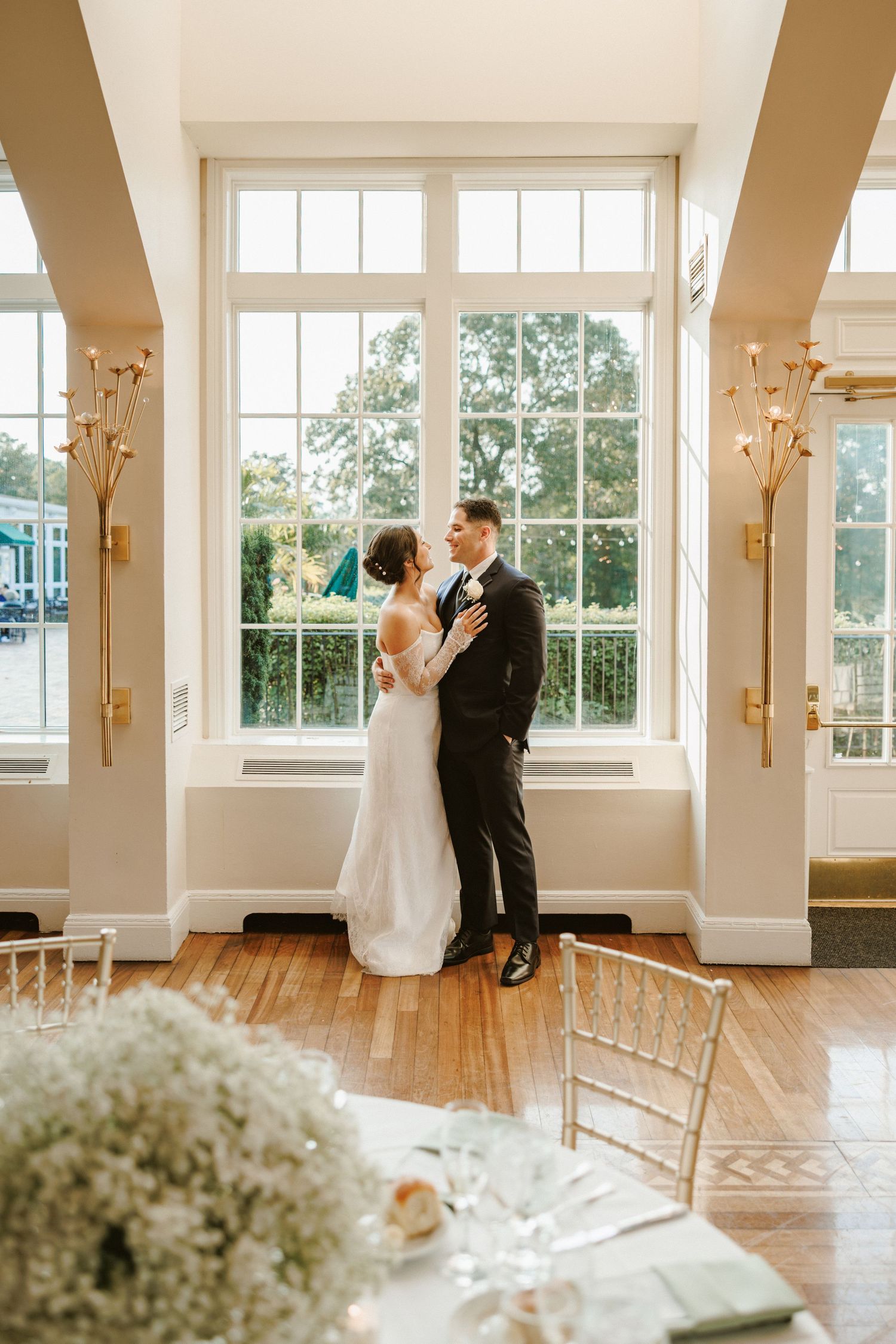 Bride and groom sharing romantic kiss in sunlit reception hall with tall windows and gold sconces.