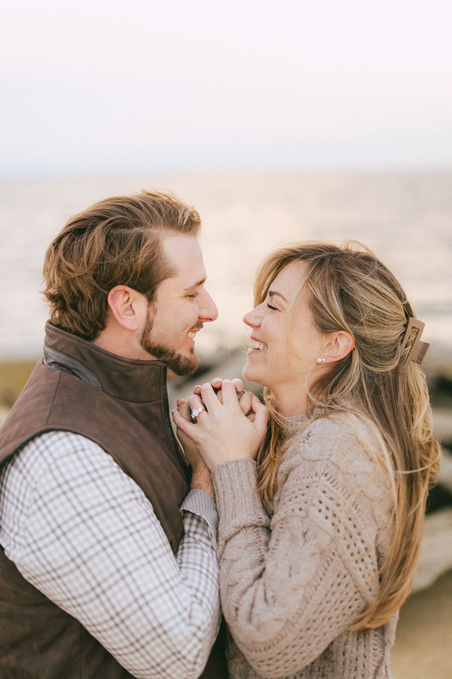 Joyful couple holds hands and laughs together in close embrace with sunlit water softly blurred behind them.