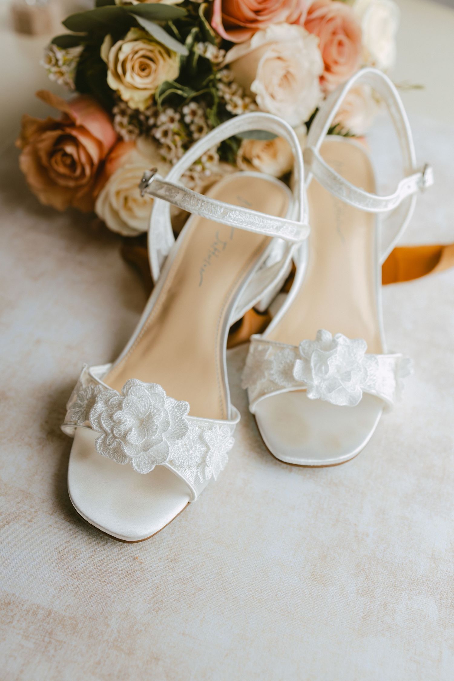 White bridal heels with lace floral details and ankle straps beside a bouquet of roses.