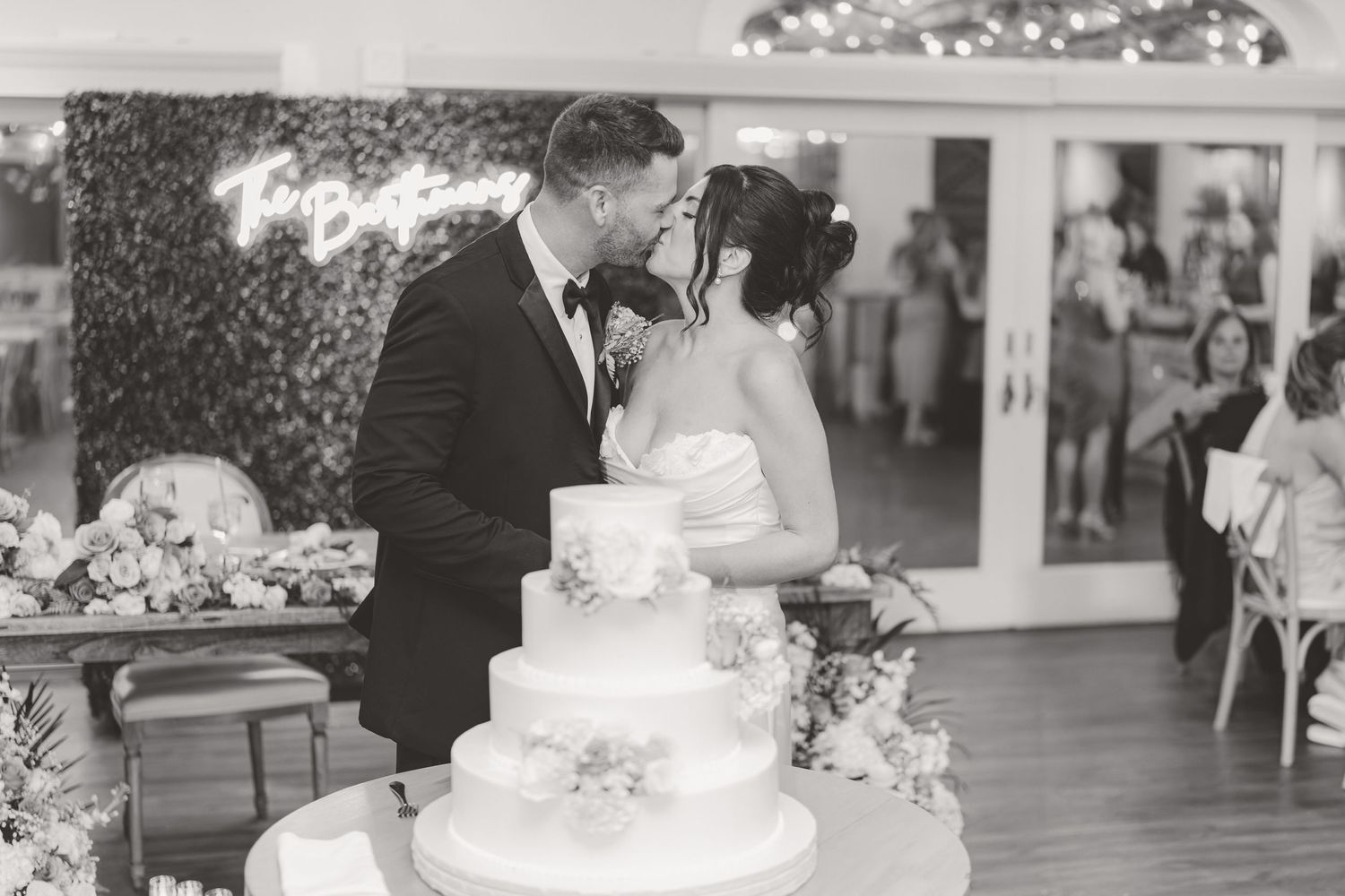 Bride and groom share a kiss behind a white tiered wedding cake at their elegant indoor reception.