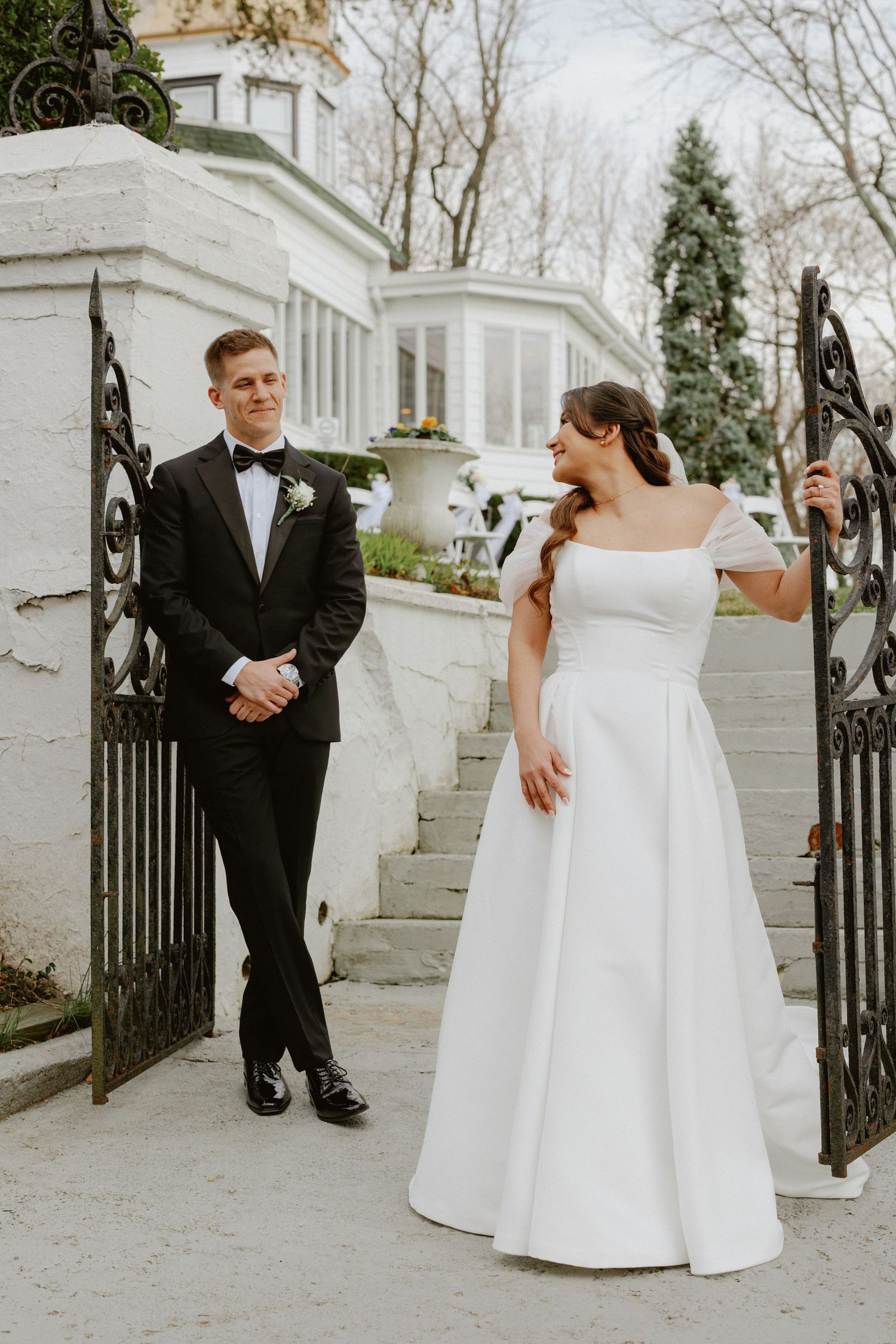 Bride in off-shoulder gown and groom in black tuxedo pose by ornate iron gates at elegant venue.