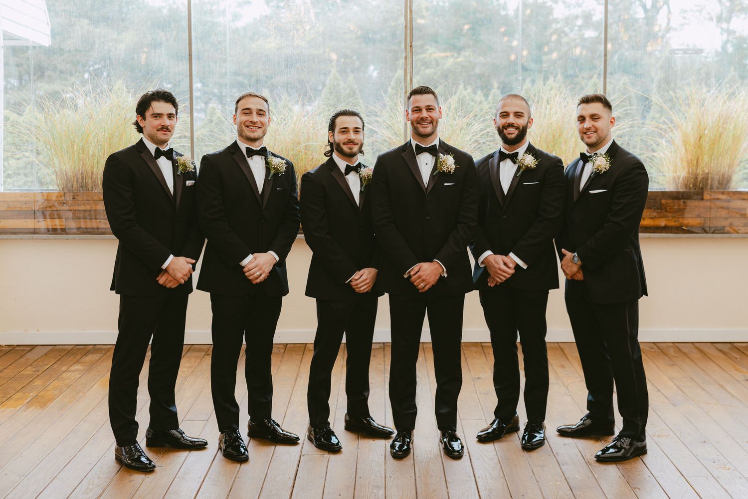 Groom and five groomsmen posing in matching black tuxedos before large windows on a wood floor.