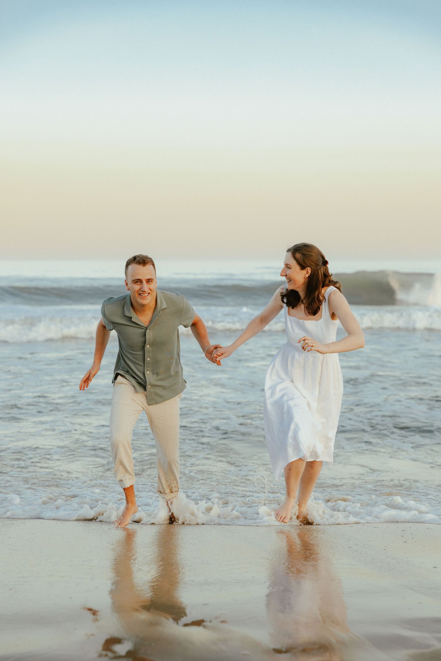 Happy couple running and holding hands through shallow ocean waves at beach during sunset.