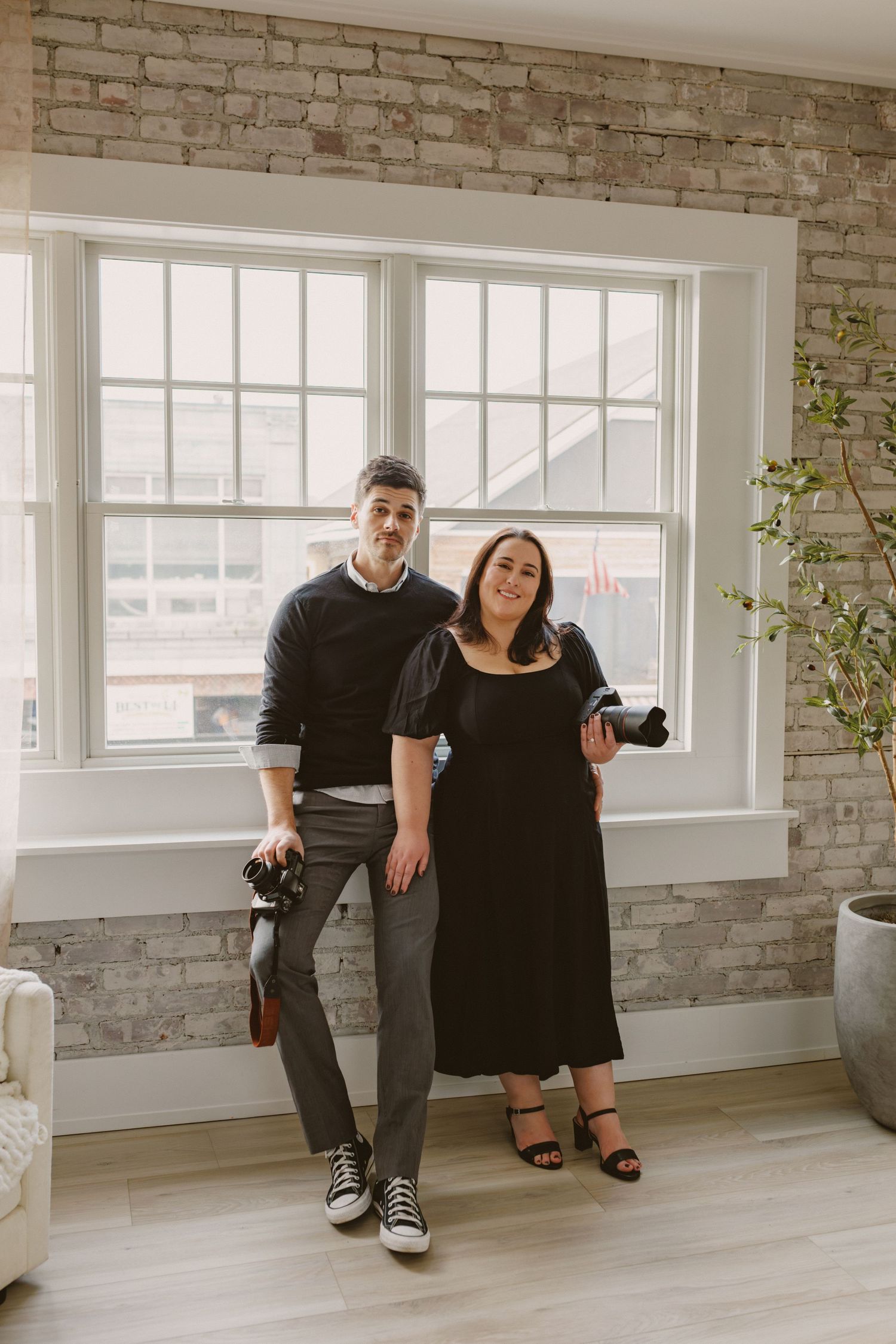 Two wedding photographers in black attire pose with cameras in bright studio with brick wall.