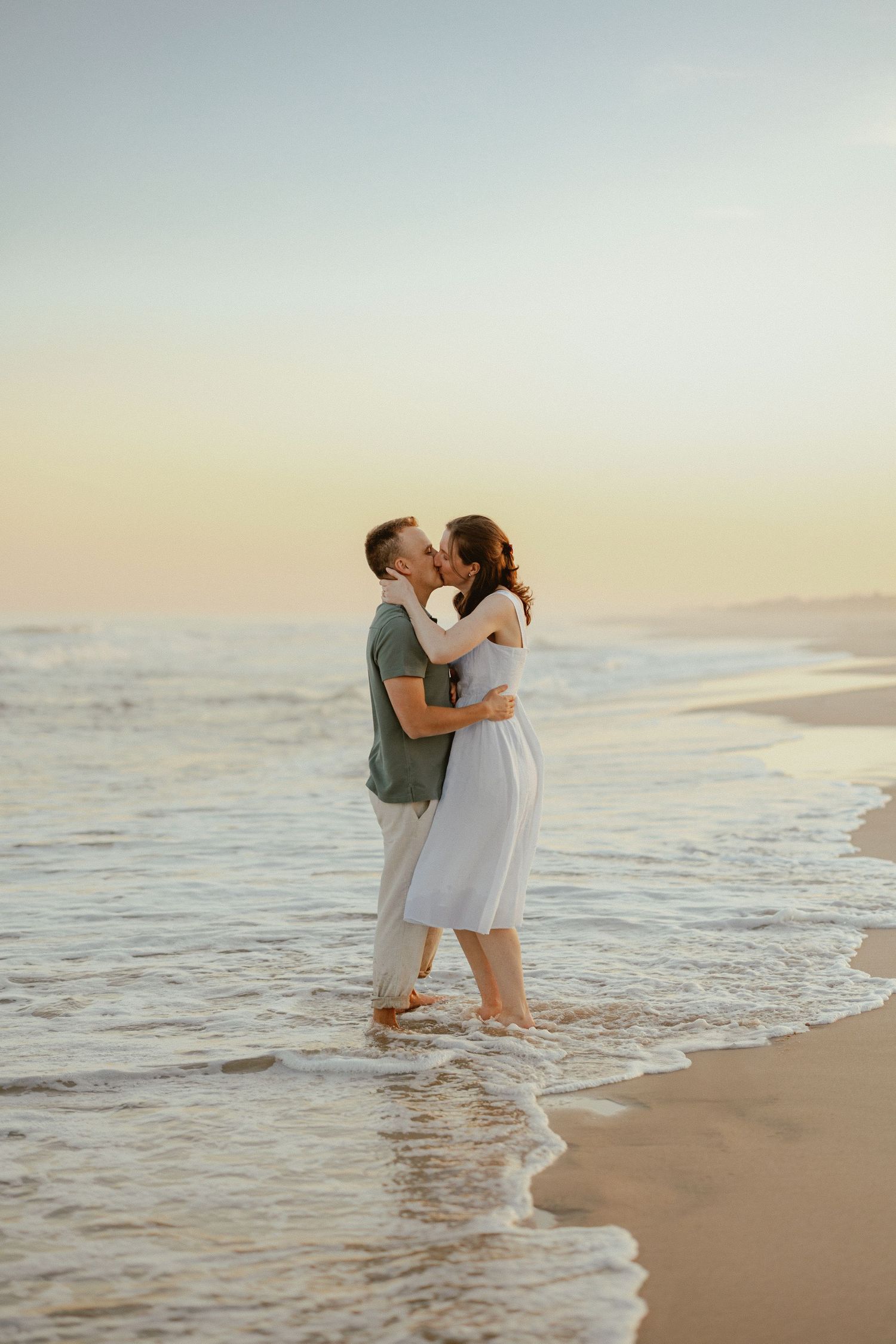 Romantic couple kissing barefoot in ocean waves at sunset on a serene sandy beach.