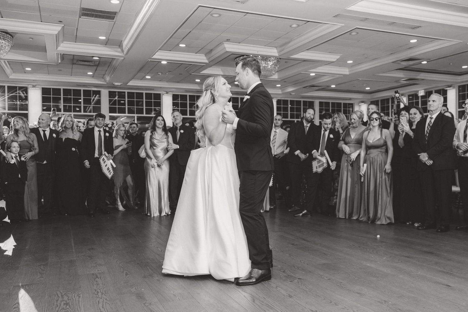 Black and white photo of bride and groom sharing their first dance surrounded by guests at the reception.