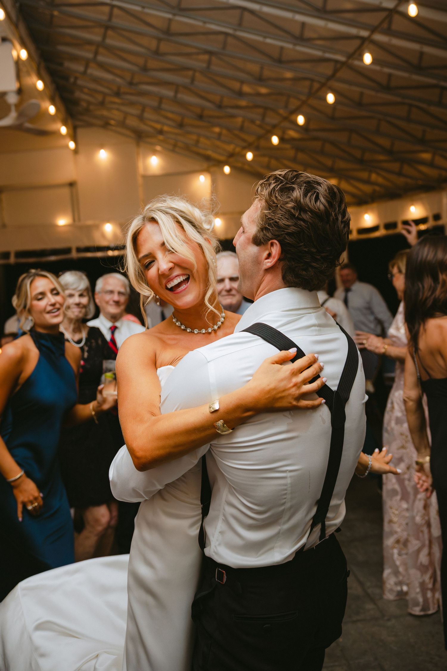 Bride laughing joyfully while dancing with groom at wedding reception under warm string lights.