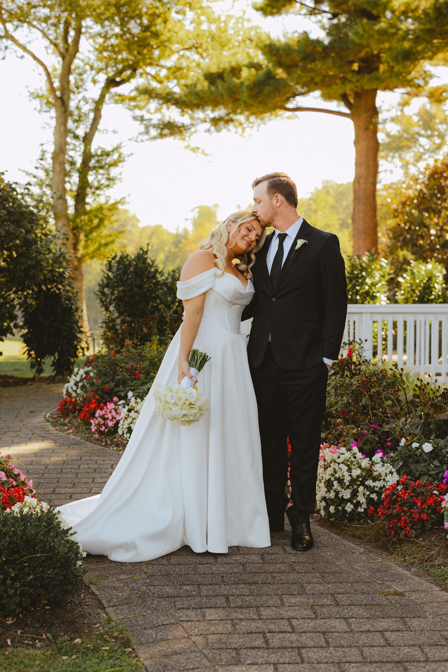 Bride and groom embrace on brick garden path surrounded by colorful flowers at golden hour.