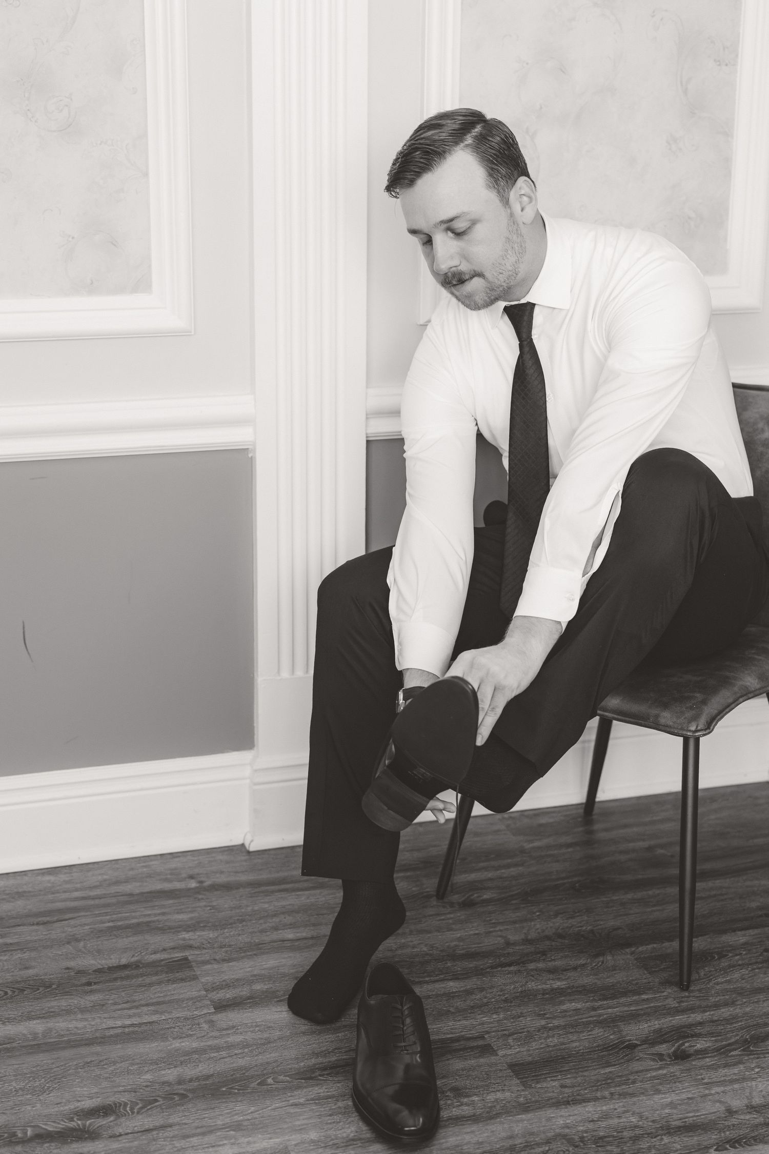 Black and white photo of groom sitting in a chair putting on dress shoes while getting ready for wedding.