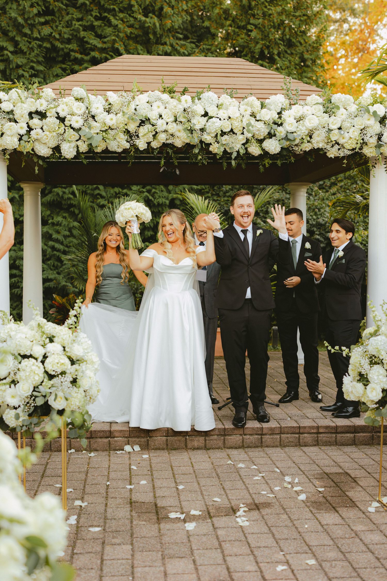 Bride and groom celebrate at outdoor gazebo ceremony adorned with white floral arrangements.