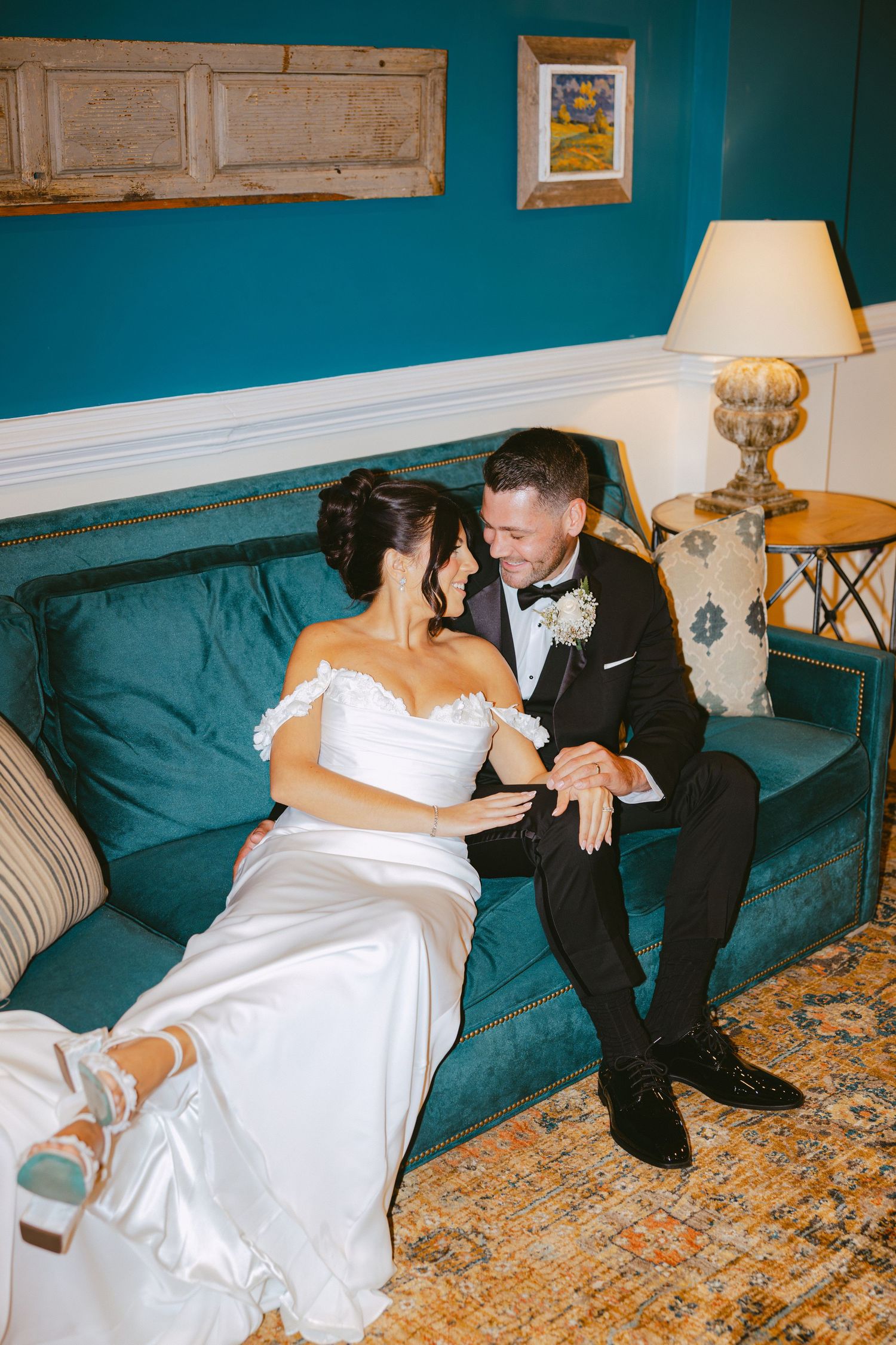 Bride and groom sitting closely on a teal velvet sofa in a richly decorated room.