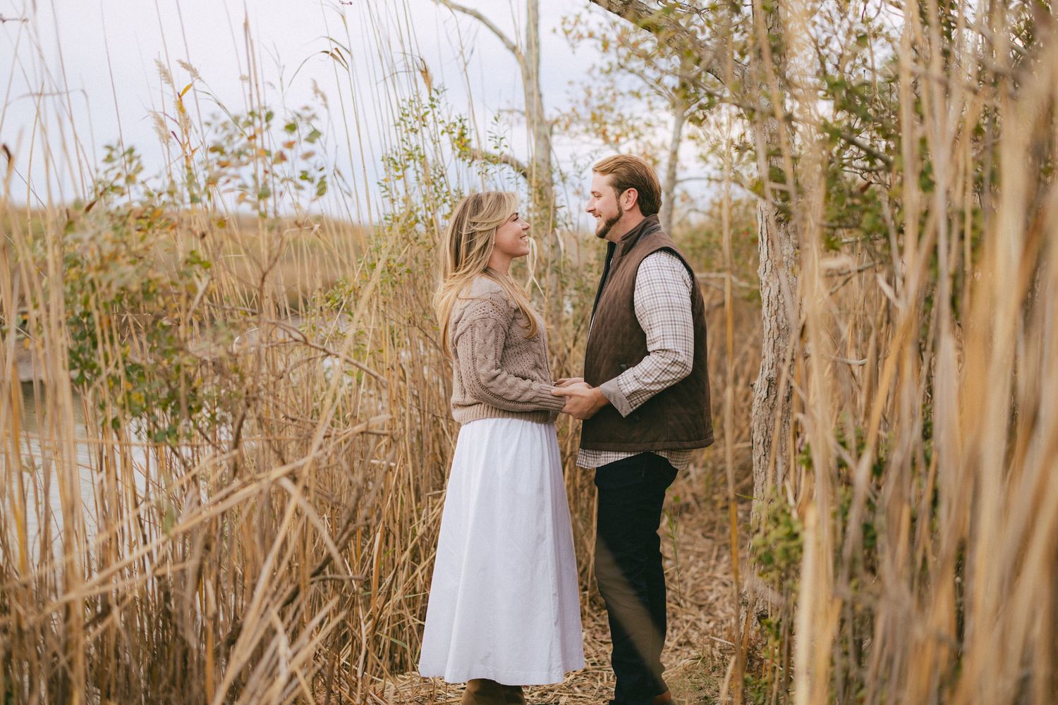 Couple holding hands and gazing at each other among tall dried reeds near a calm waterway.