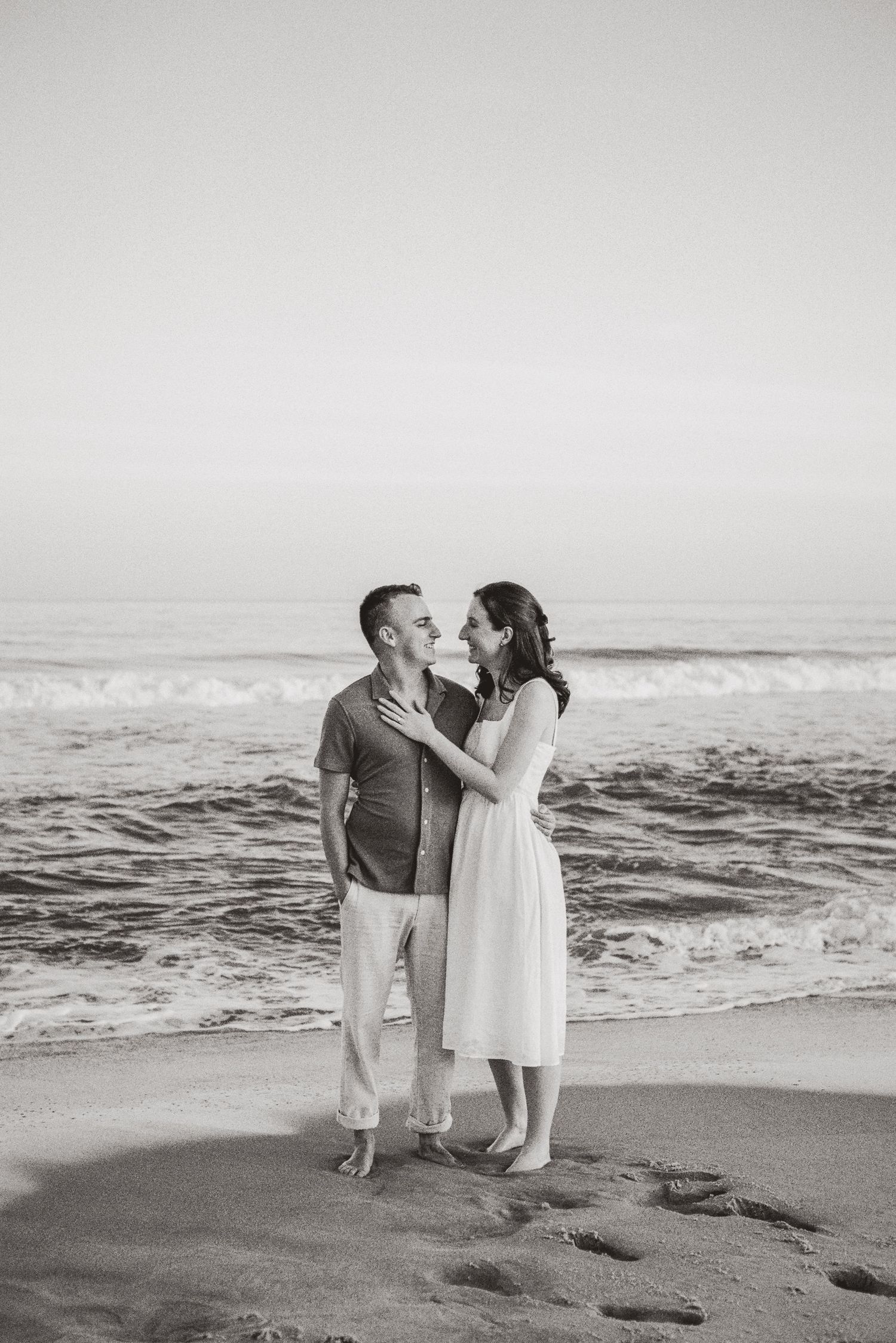 Black and white image of a romantic couple nearly kissing on a beach with ocean waves in the background.