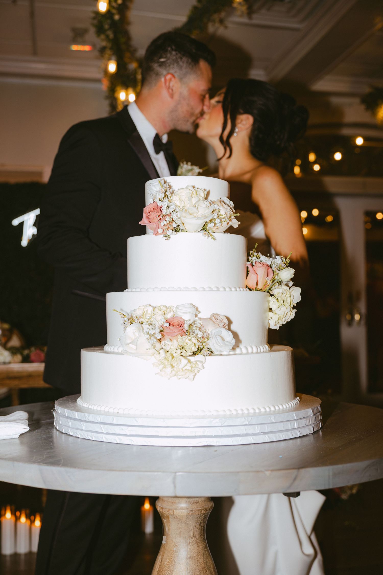 Couple kisses behind a beautiful four-tier white wedding cake adorned with pink flowers.