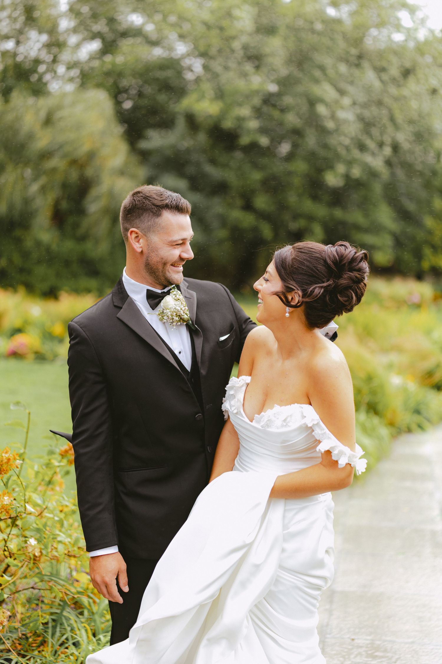 Bride and groom laughing together while walking along a garden path with colorful flowers.