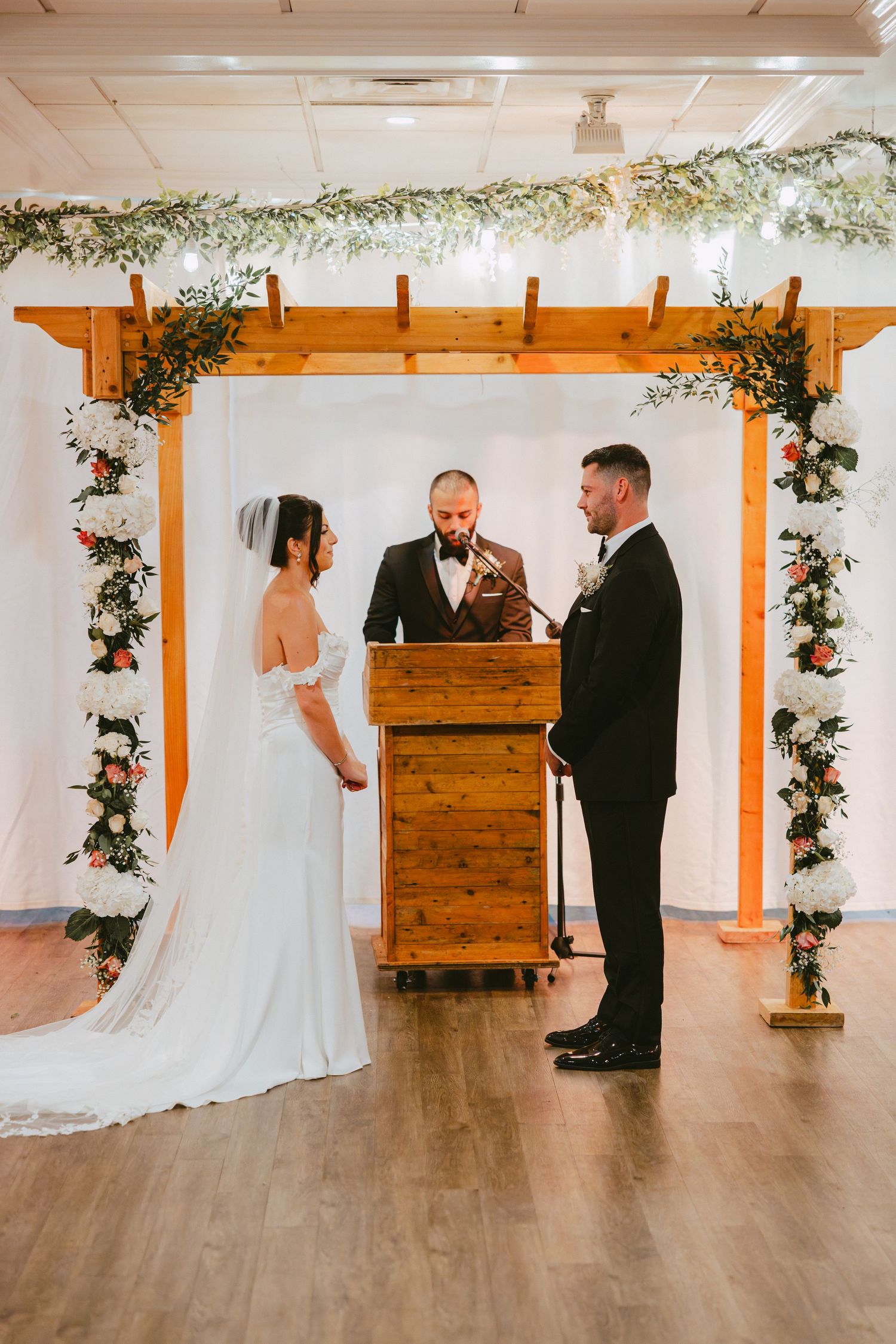Bride and groom exchange vows under floral wooden arch with officiant at indoor wedding ceremony.