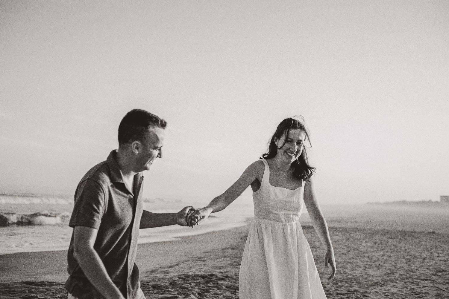 Black and white photo of a happy couple holding hands and walking along a sunny beach shoreline.