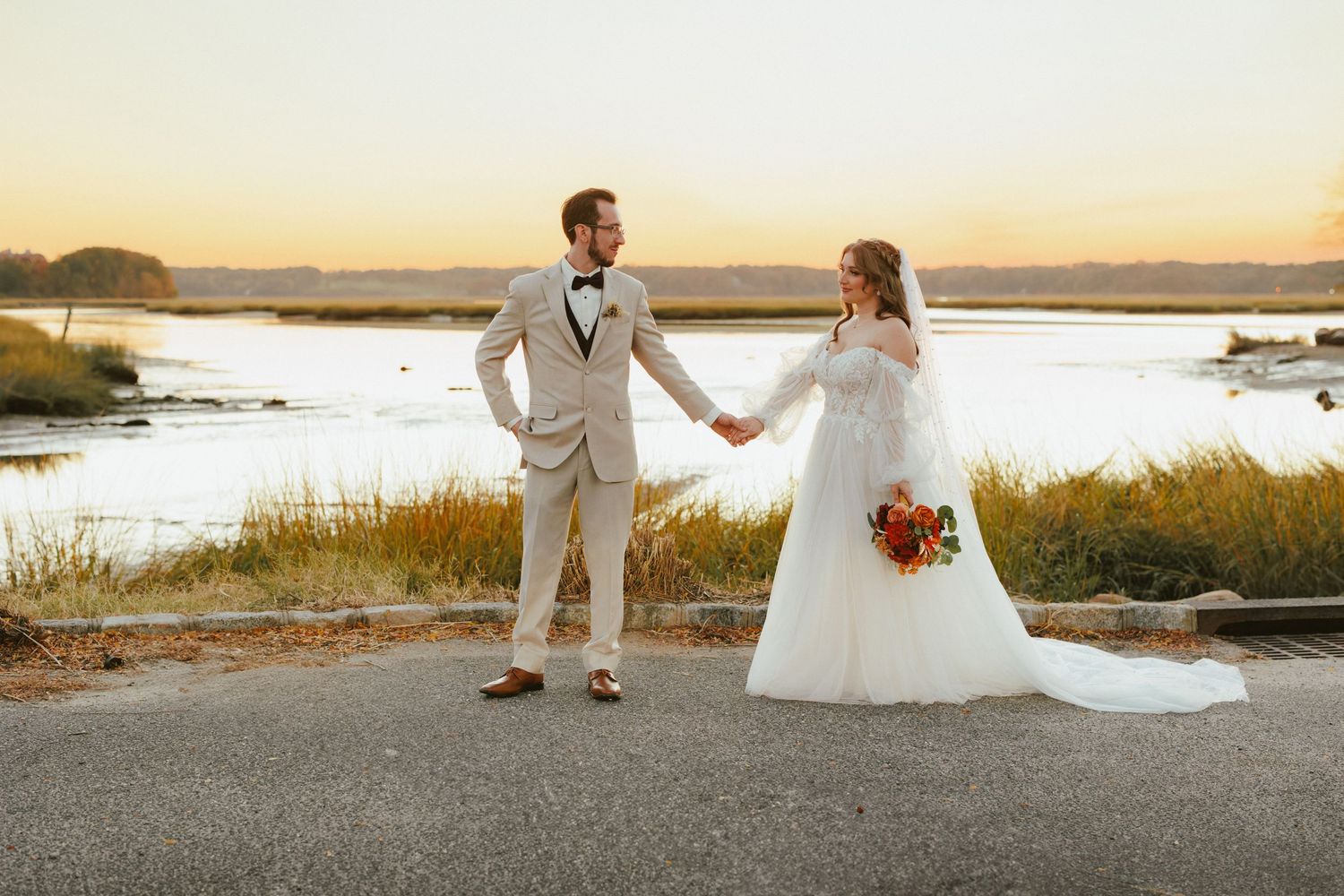 Bride and groom hold hands by serene waterway at sunset, bride in lace gown, groom in tan suit.