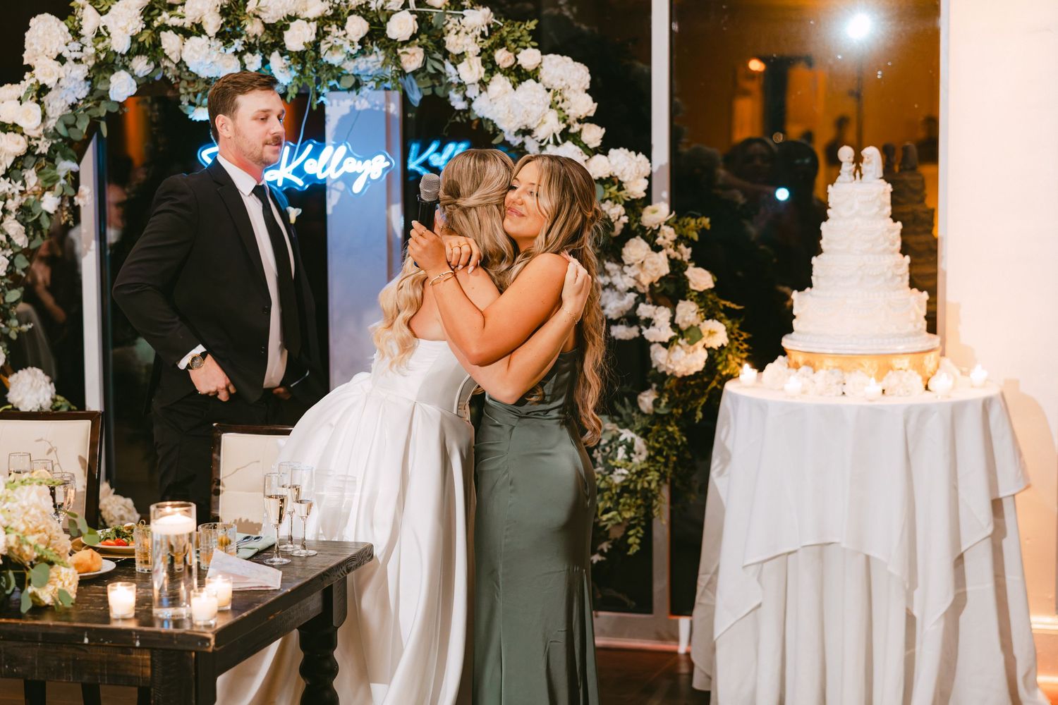 Bride and bridesmaid share an emotional hug at wedding reception with white floral arrangements and neon sign.