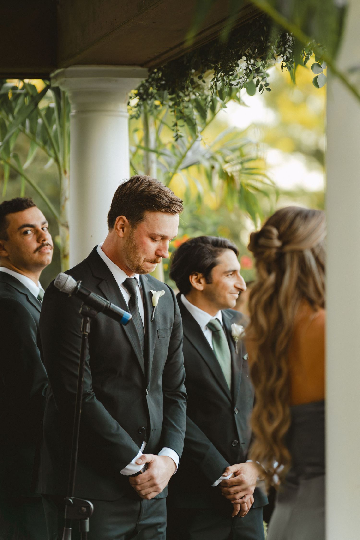 Groom standing at altar in black suit watching bride approach during outdoor wedding ceremony.