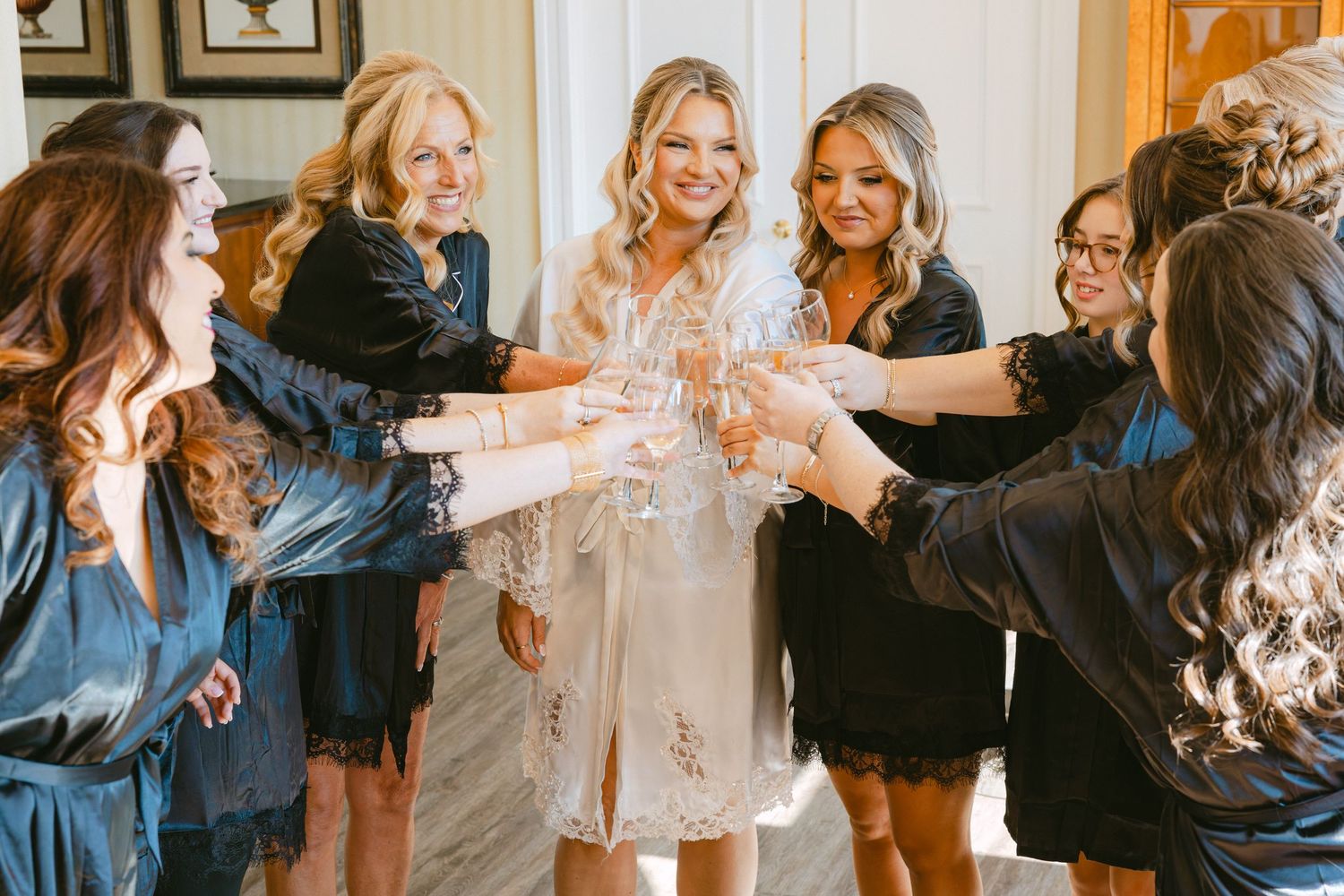 Bride in lace robe toasting champagne with bridesmaids in navy robes during wedding morning preparations.
