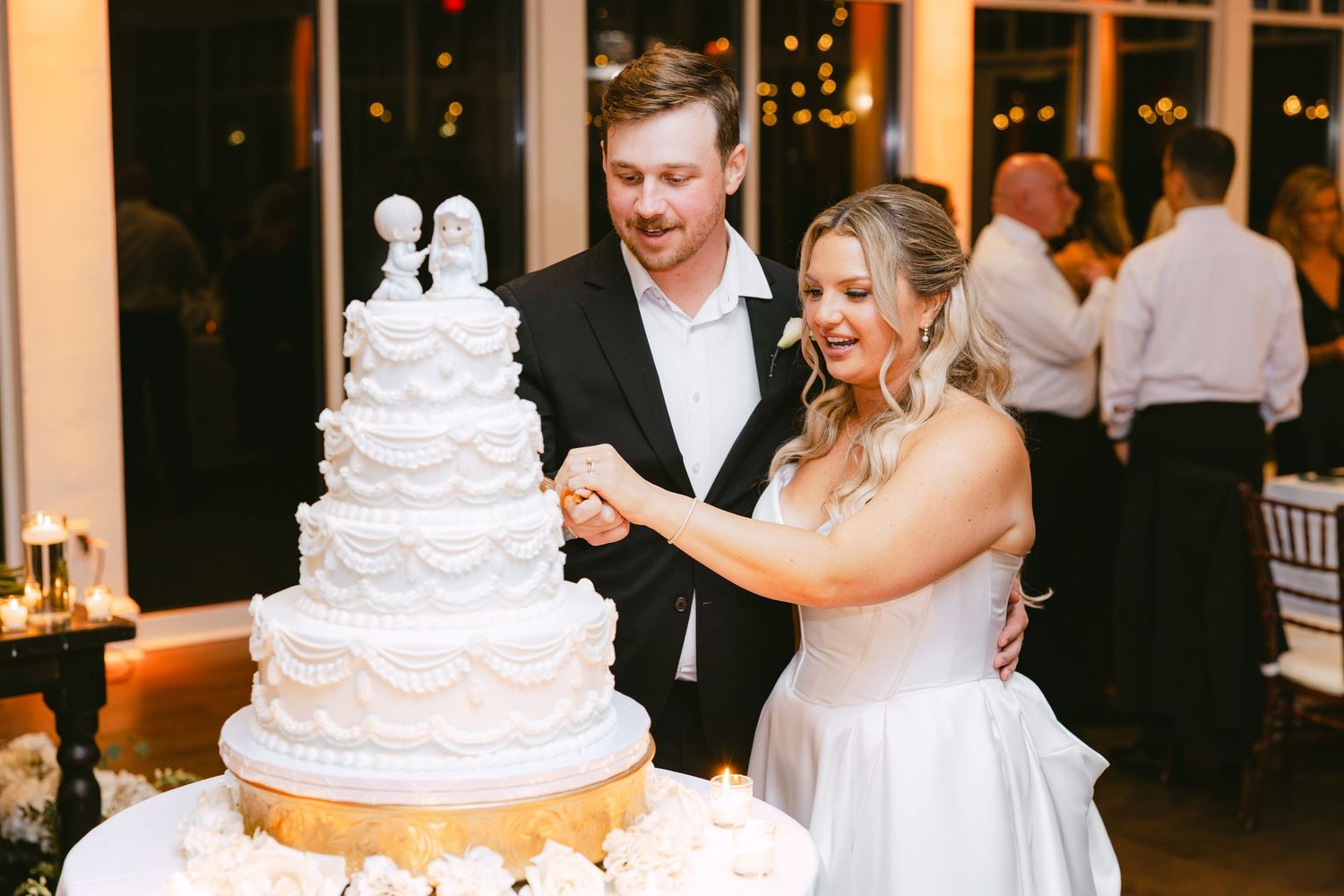 Bride and groom cutting their elegant four-tier white wedding cake topped with figurines at their reception.