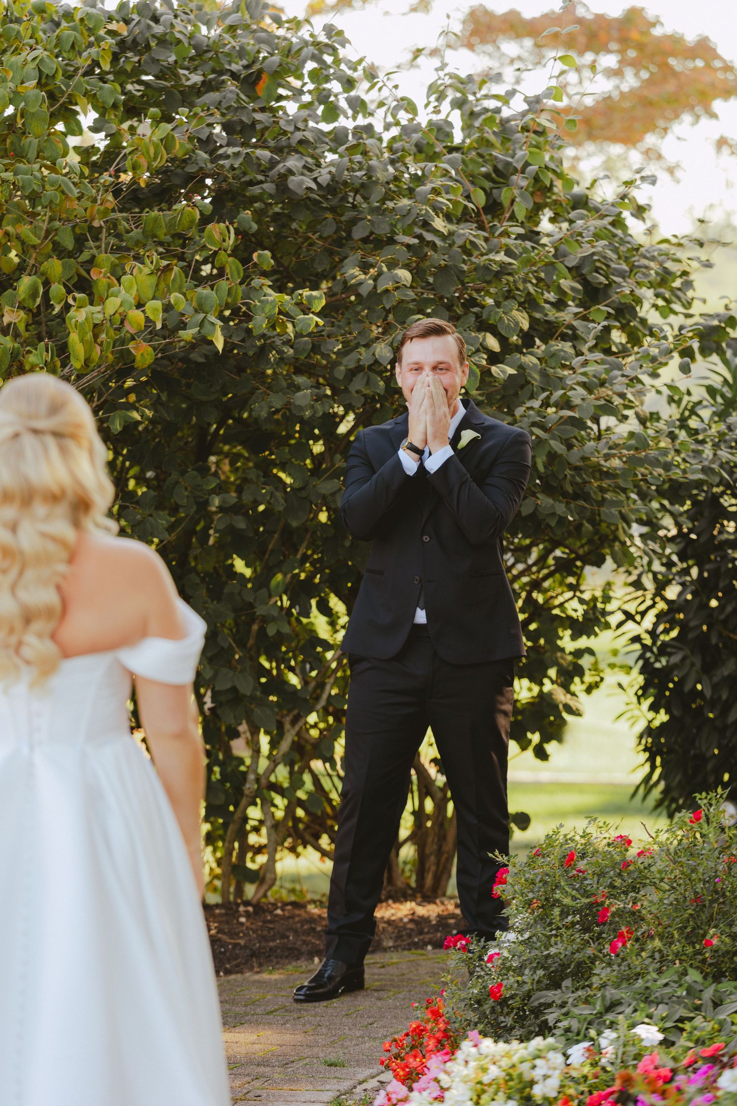 Groom covers his mouth in emotional awe during first look with bride in outdoor garden setting.