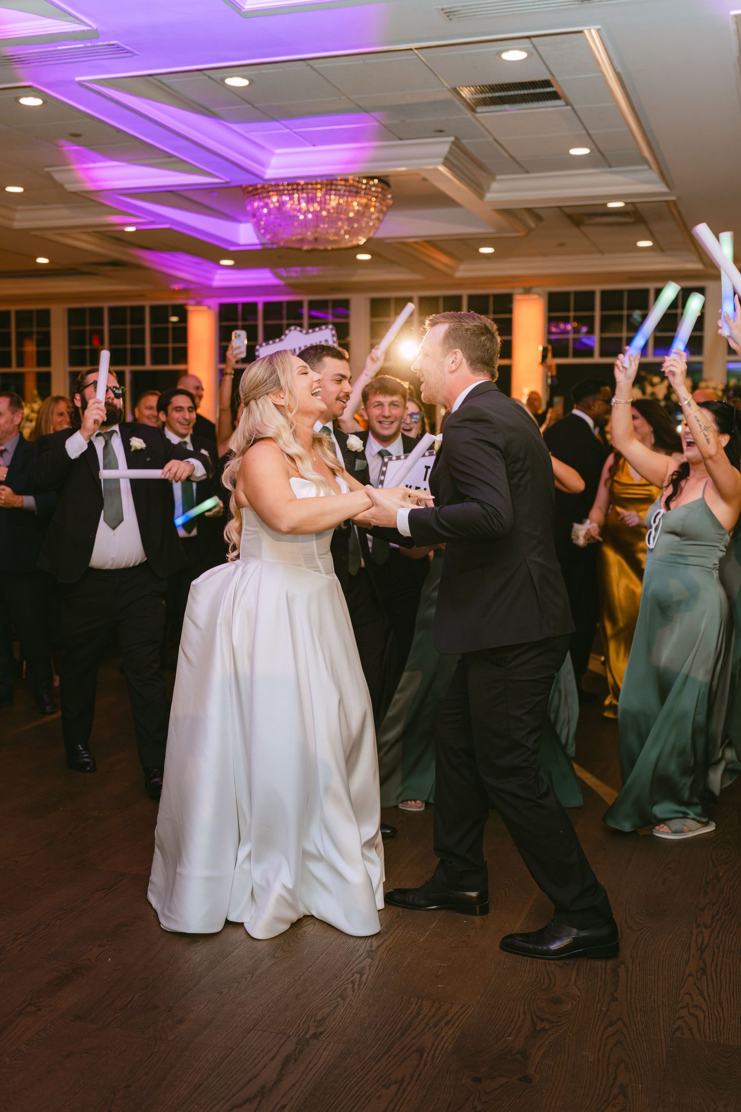 Bride and groom share a first dance surrounded by guests waving glow sticks at wedding reception.
