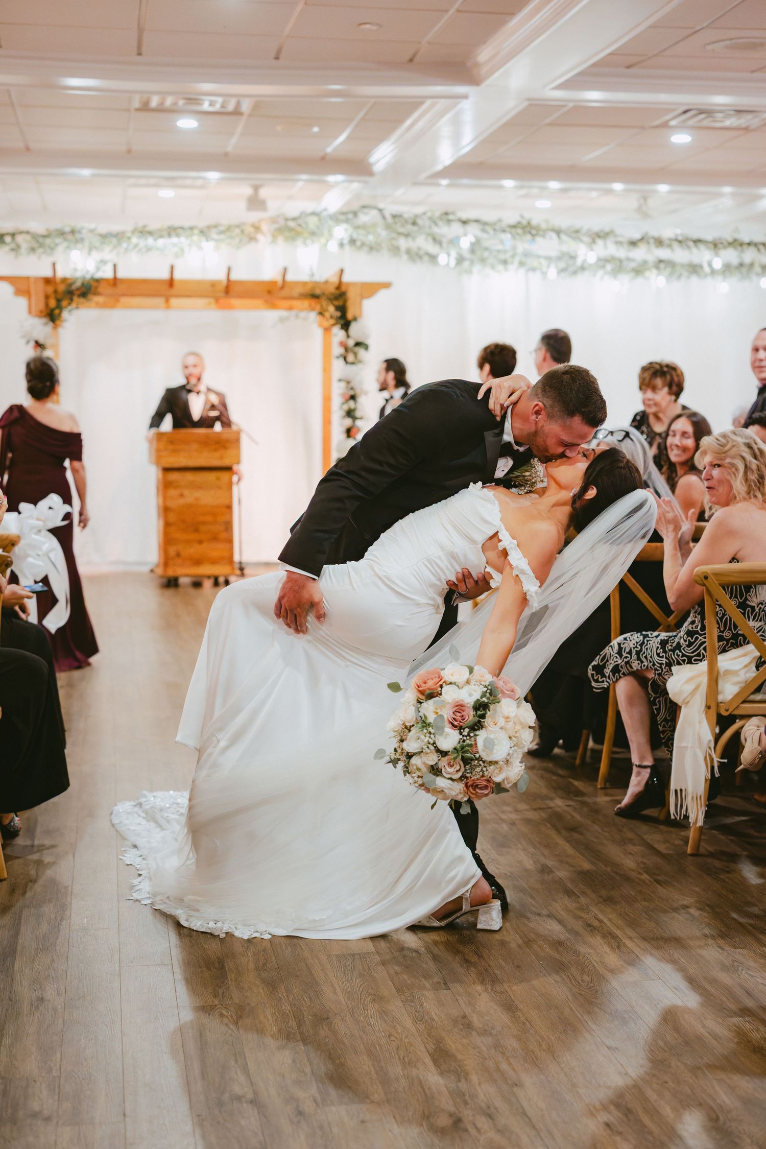 Groom dips bride for romantic kiss while she holds bouquet after indoor wedding ceremony.
