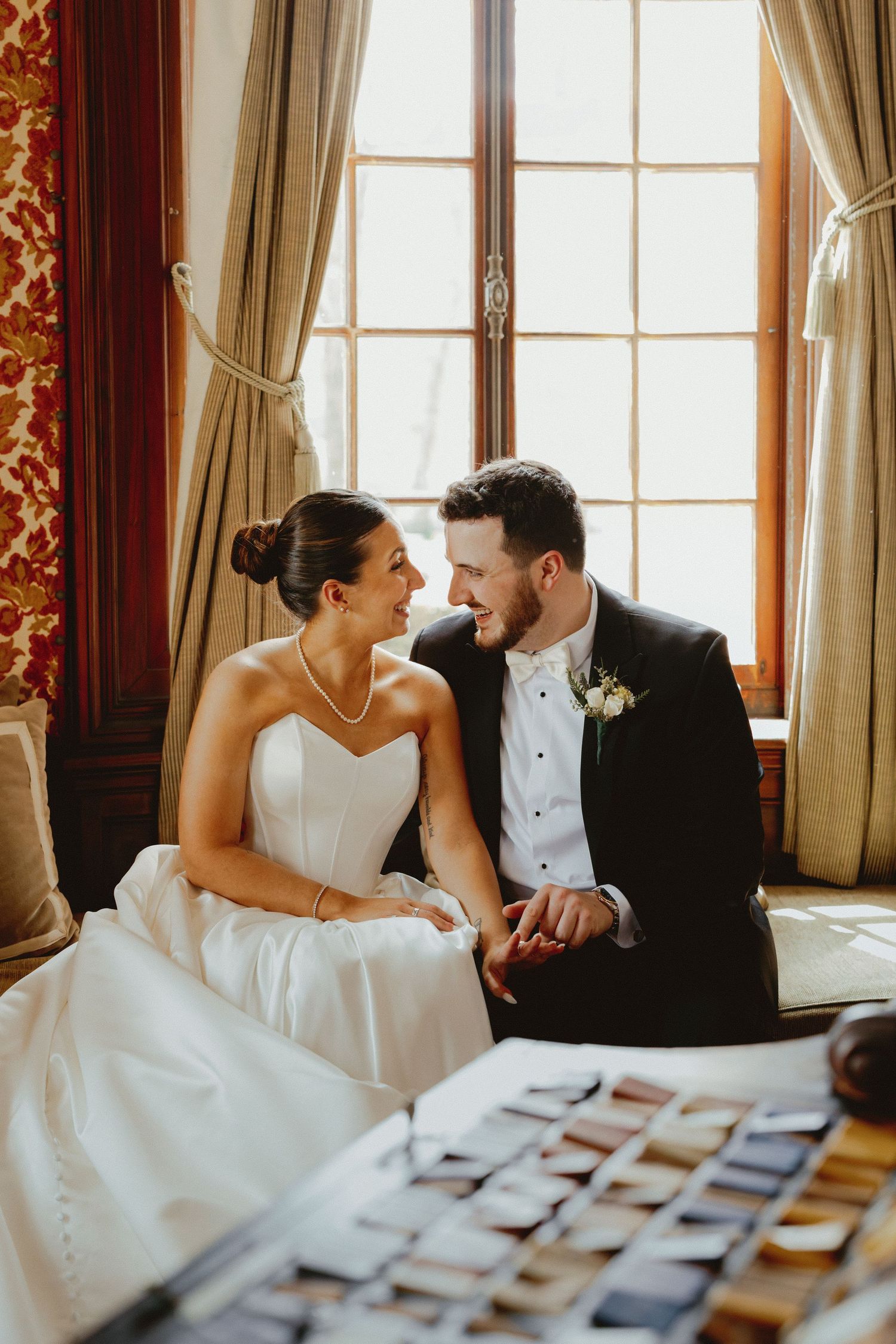 Bride and groom sit together holding hands by sunlit window in ornate room on wedding day.